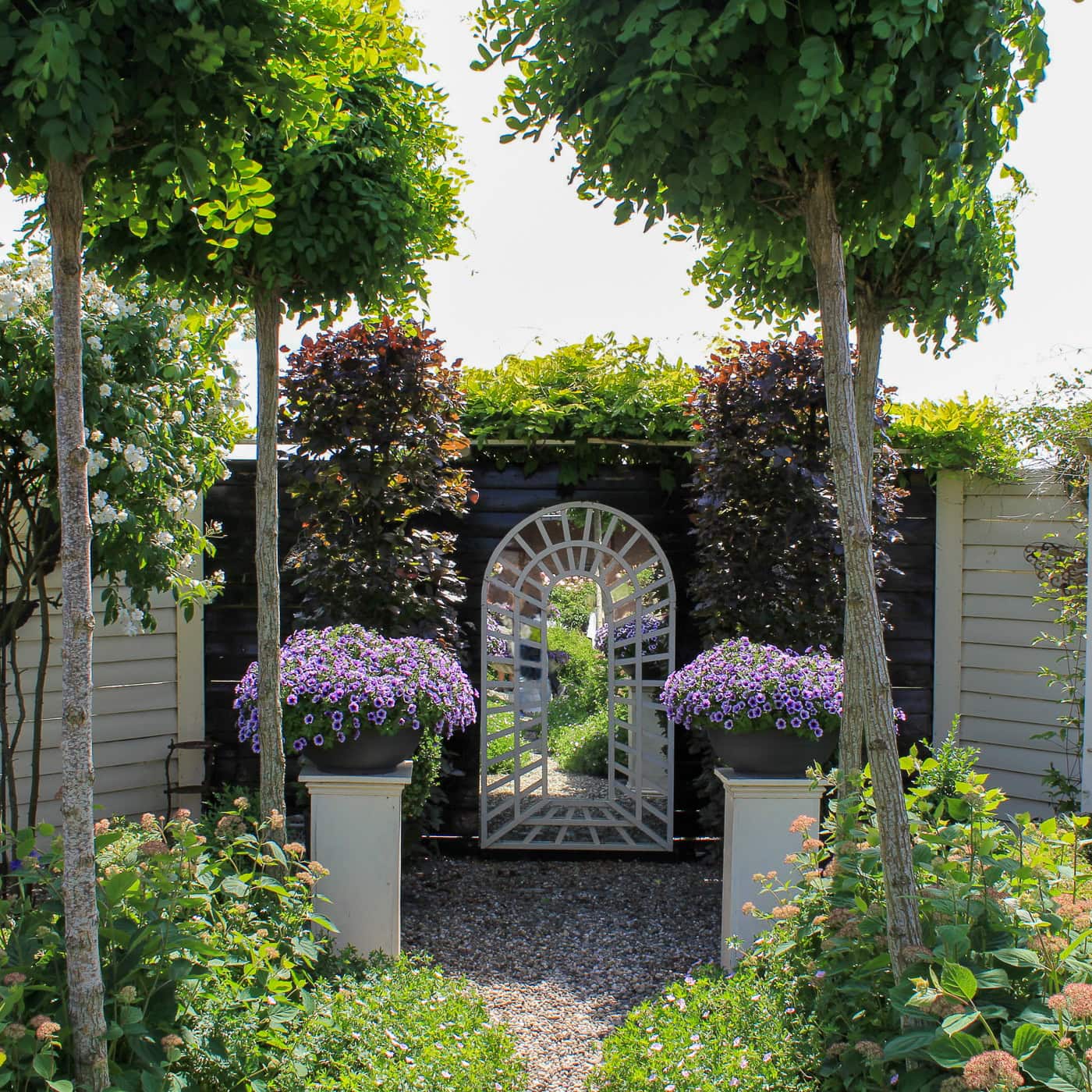 A garden with manicured trees, flowering plants, and two large pots of purple flowers flanking an arched gate set in a wooden fence.