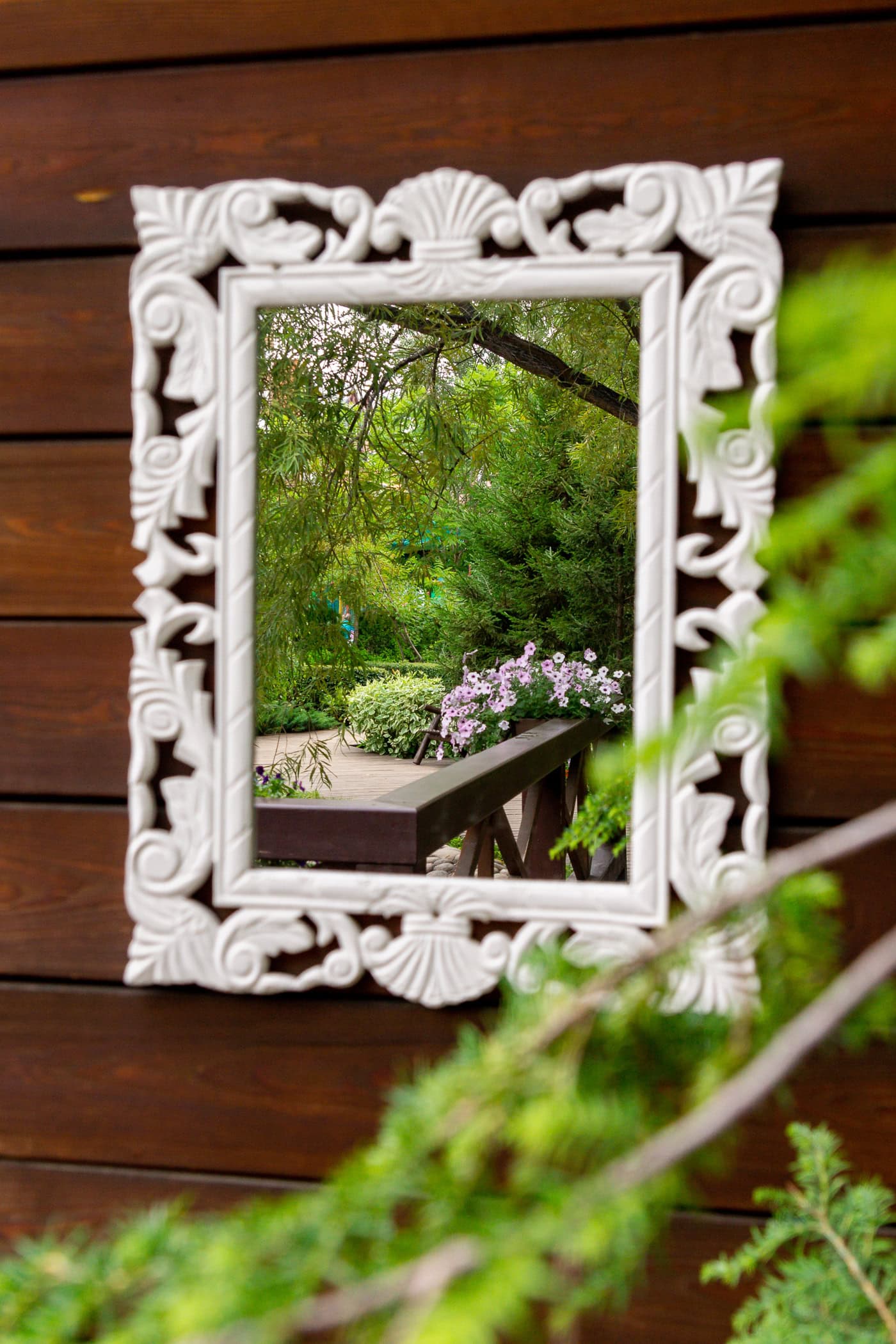 A decorative white-framed mirror is mounted on a wooden wall, reflecting a lush garden scene with green trees and pink flowers.
