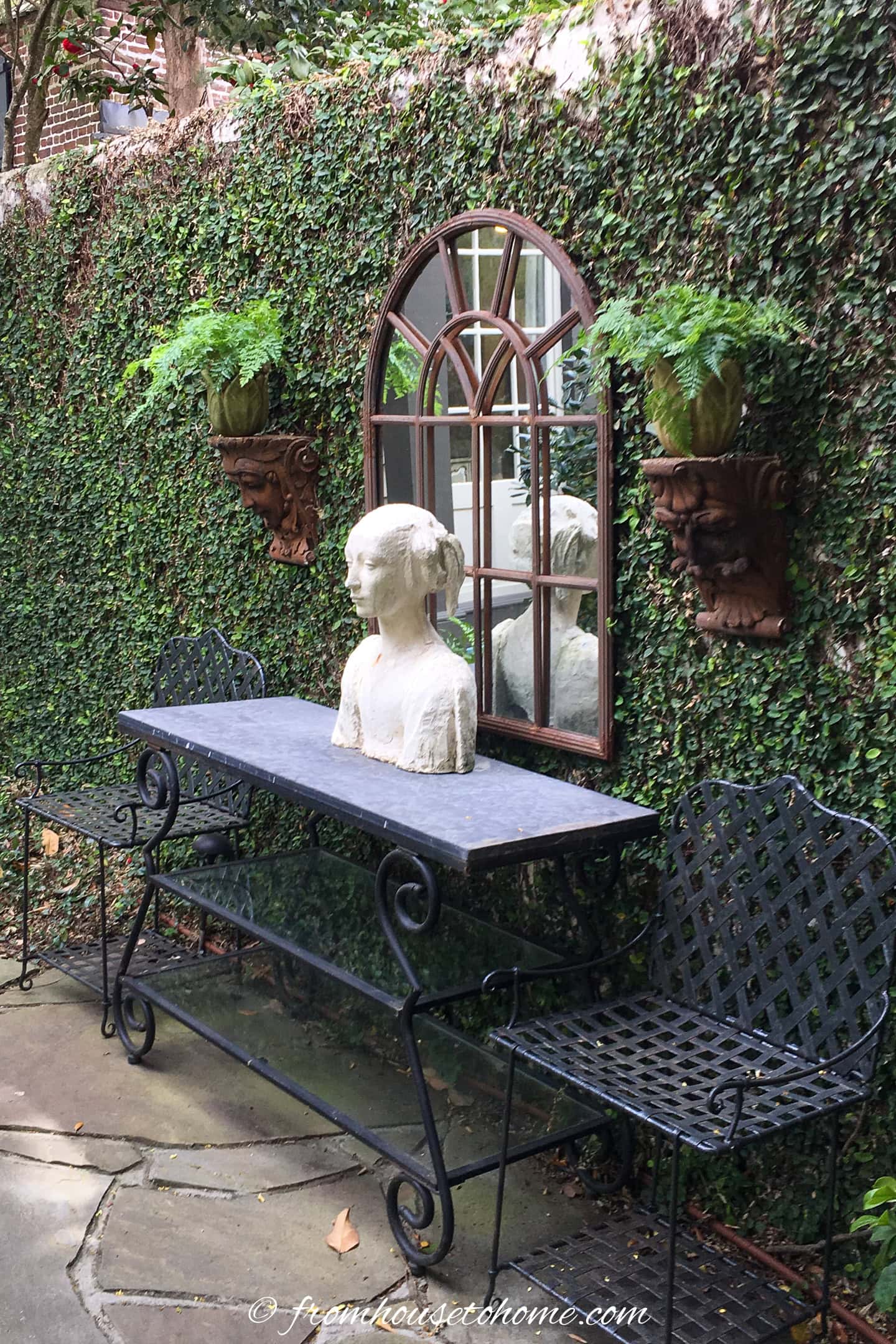 A stone bust sits on a black metal table between two benches in a garden, with a mirror and two potted ferns mounted on an ivy-covered wall behind it.