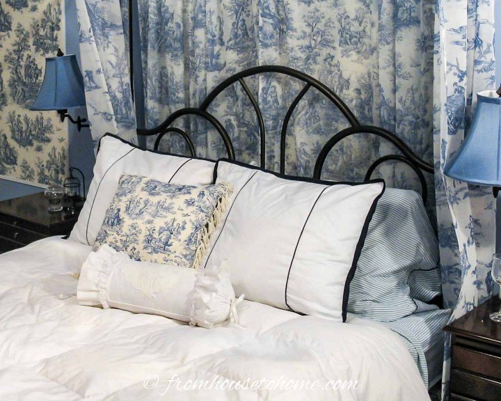 Blue and white striped sheets with a white duvet and toile throw pillows on a wrought iron bed