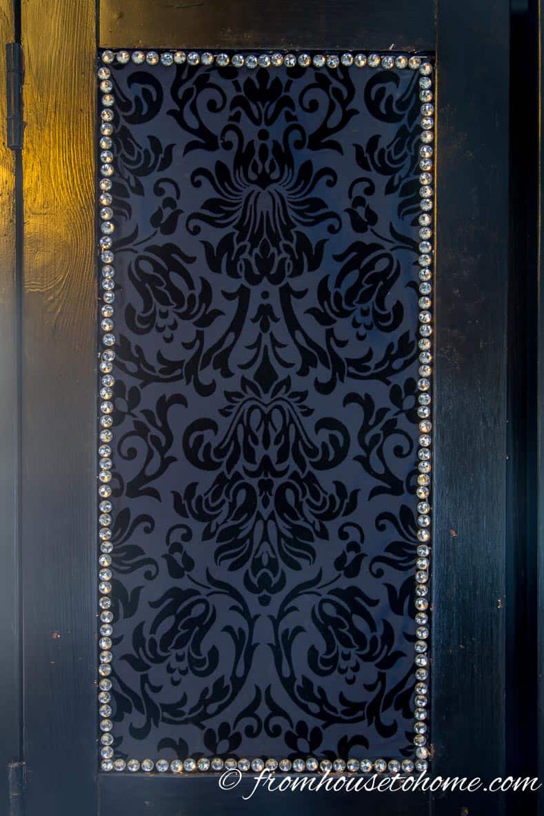 Black armoire door panel covered with fabric surrounded by crystal upholstery tacks