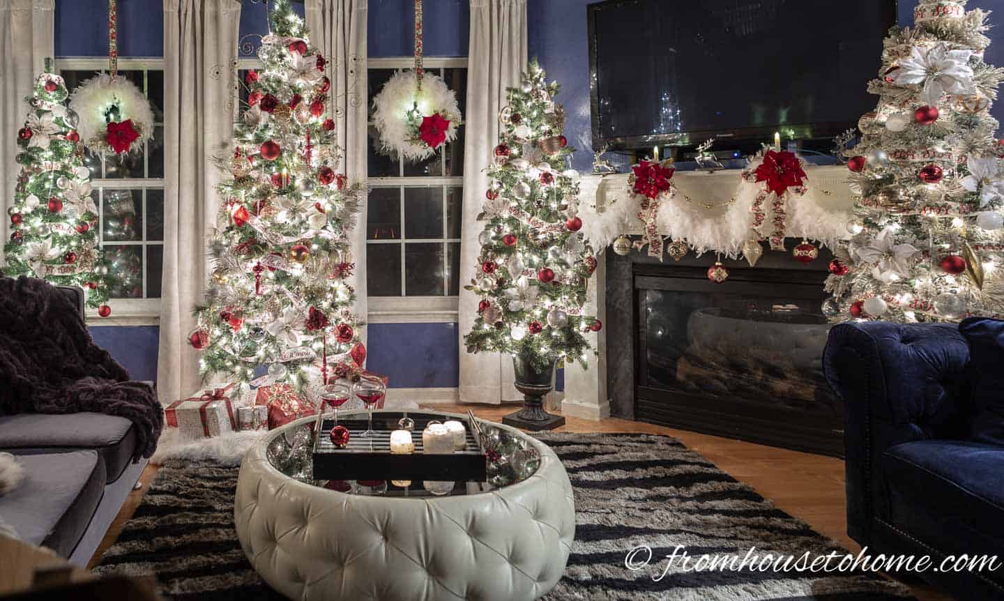 A living room with 4 Christmas trees decorated in red, white and gold Christmas decorations, 2 white wreaths on the windows and a white garland over the fireplace