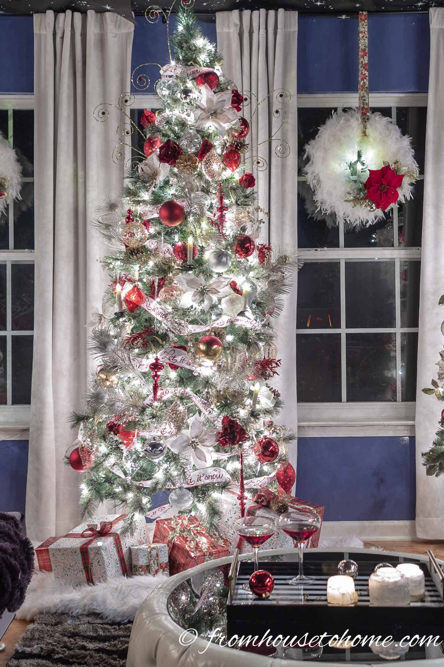 A Christmas tree decorated with red, white and gold Christmas ornaments in front of a window with a white wreath