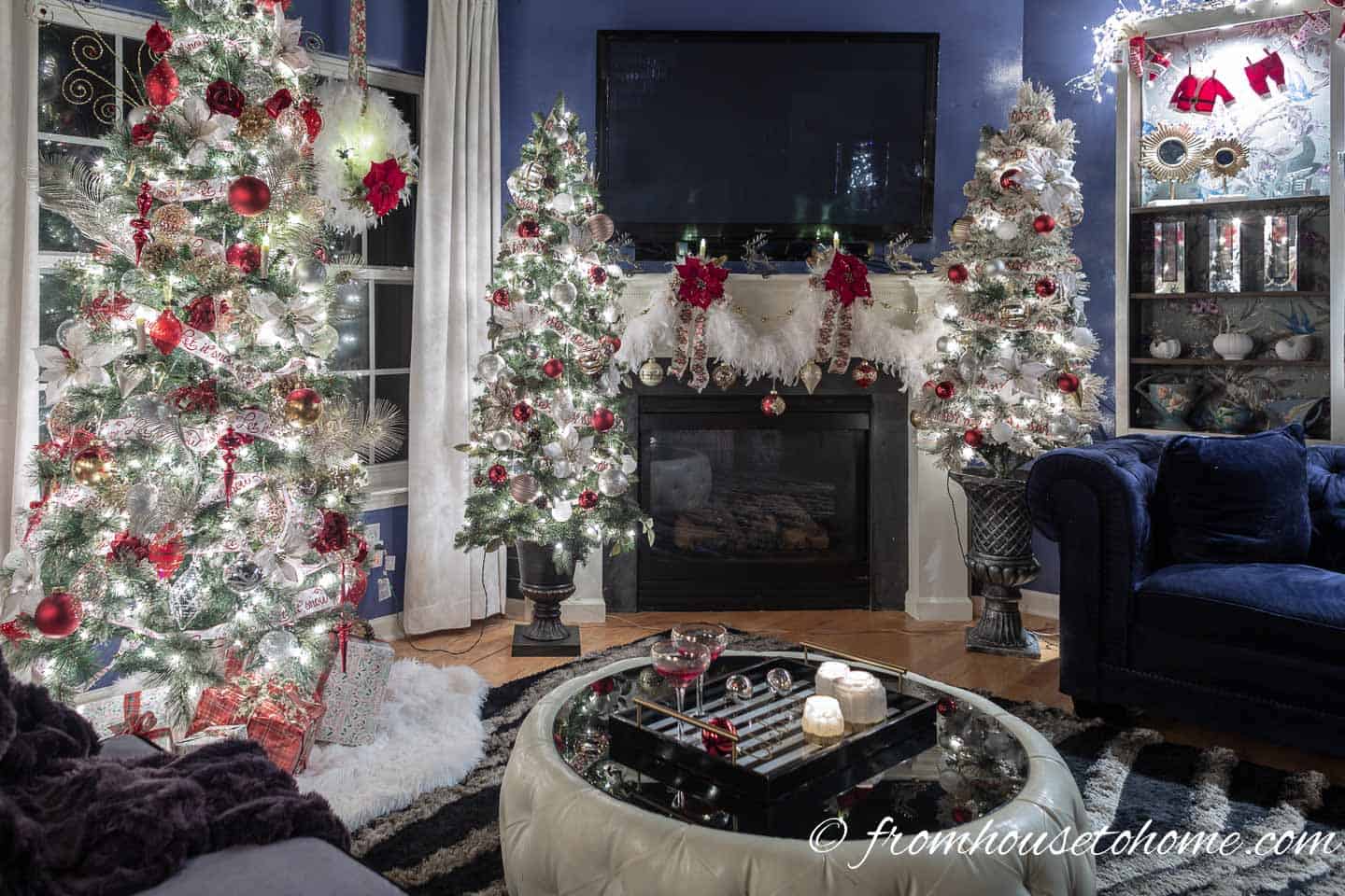Living room with 3 Christmas trees and a fireplace decorated with red, white and gold Christmas decorations