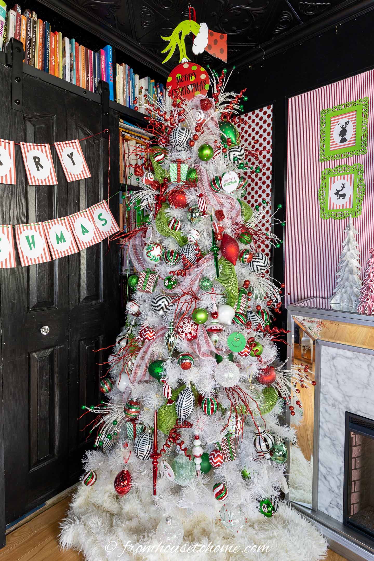 A white Christmas tree decorated with red, green, and white ornaments stands in a festive room with striped walls, a bookshelf, and a faux fireplace.