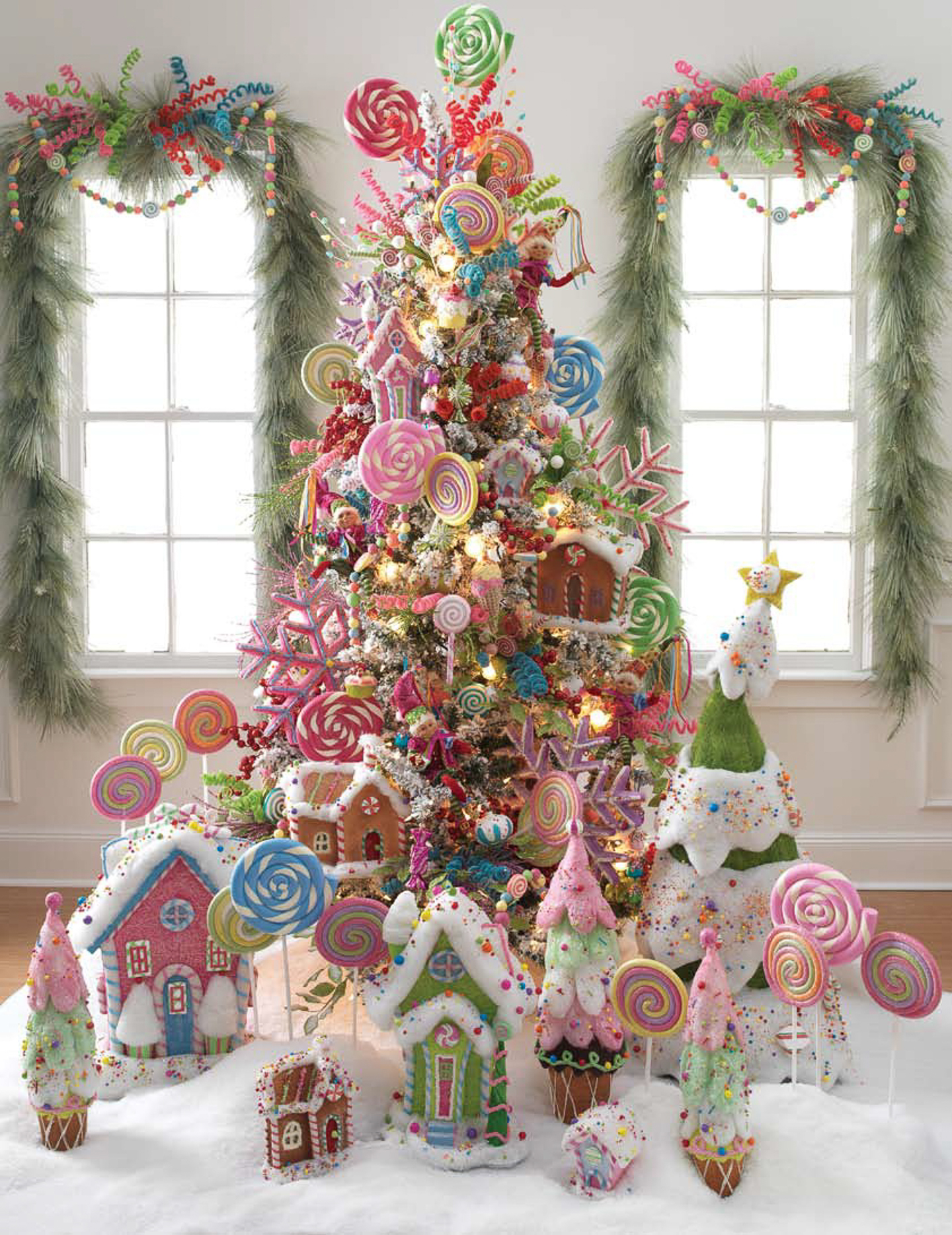 A Christmas tree decorated with colorful candy-themed ornaments, surrounded by gingerbread houses and large lollipop props, set in a festive room with garland on the windows.