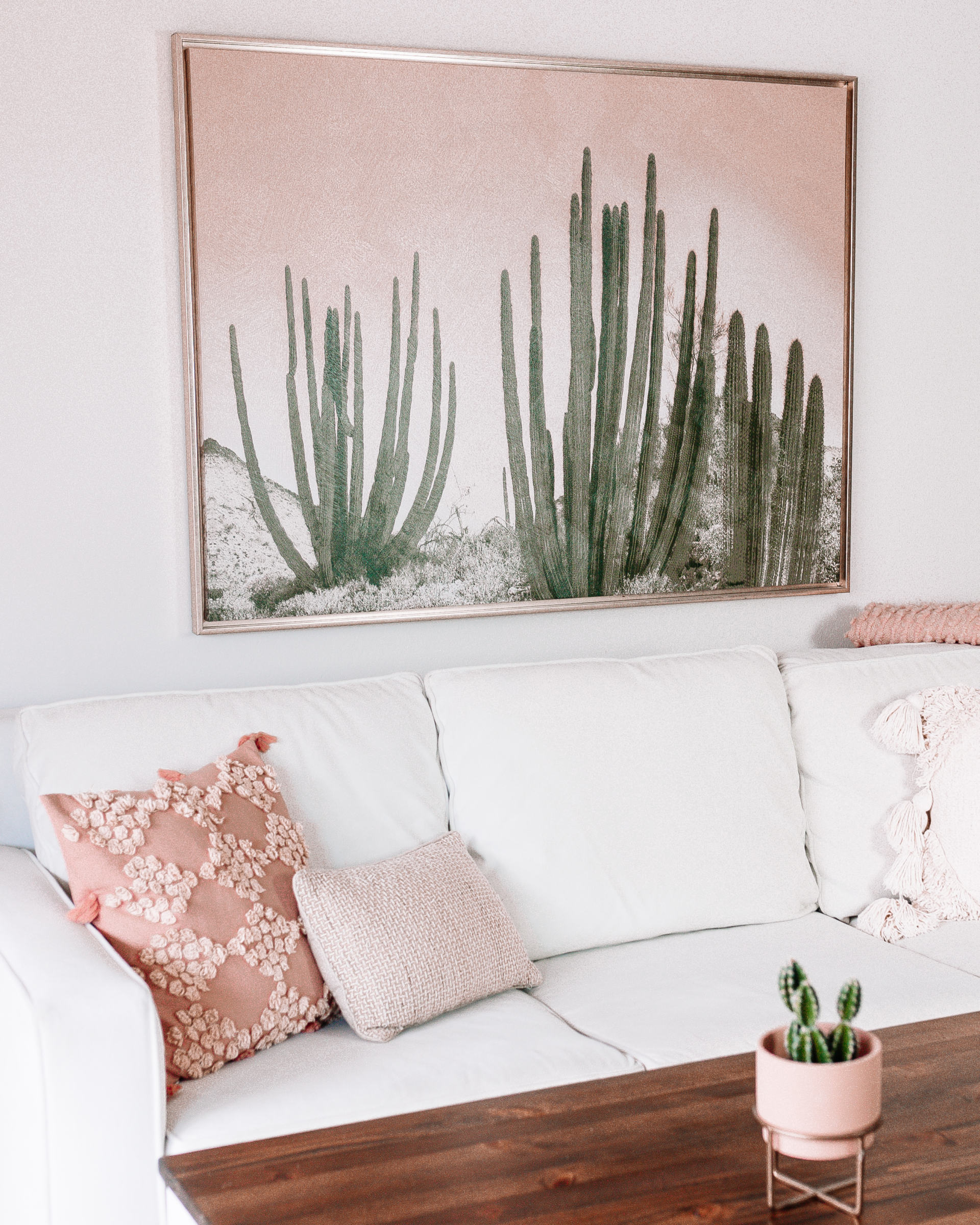 A white sofa with pink and textured pillows sits below a large framed cactus artwork; a small potted cactus is on a wooden coffee table in front.