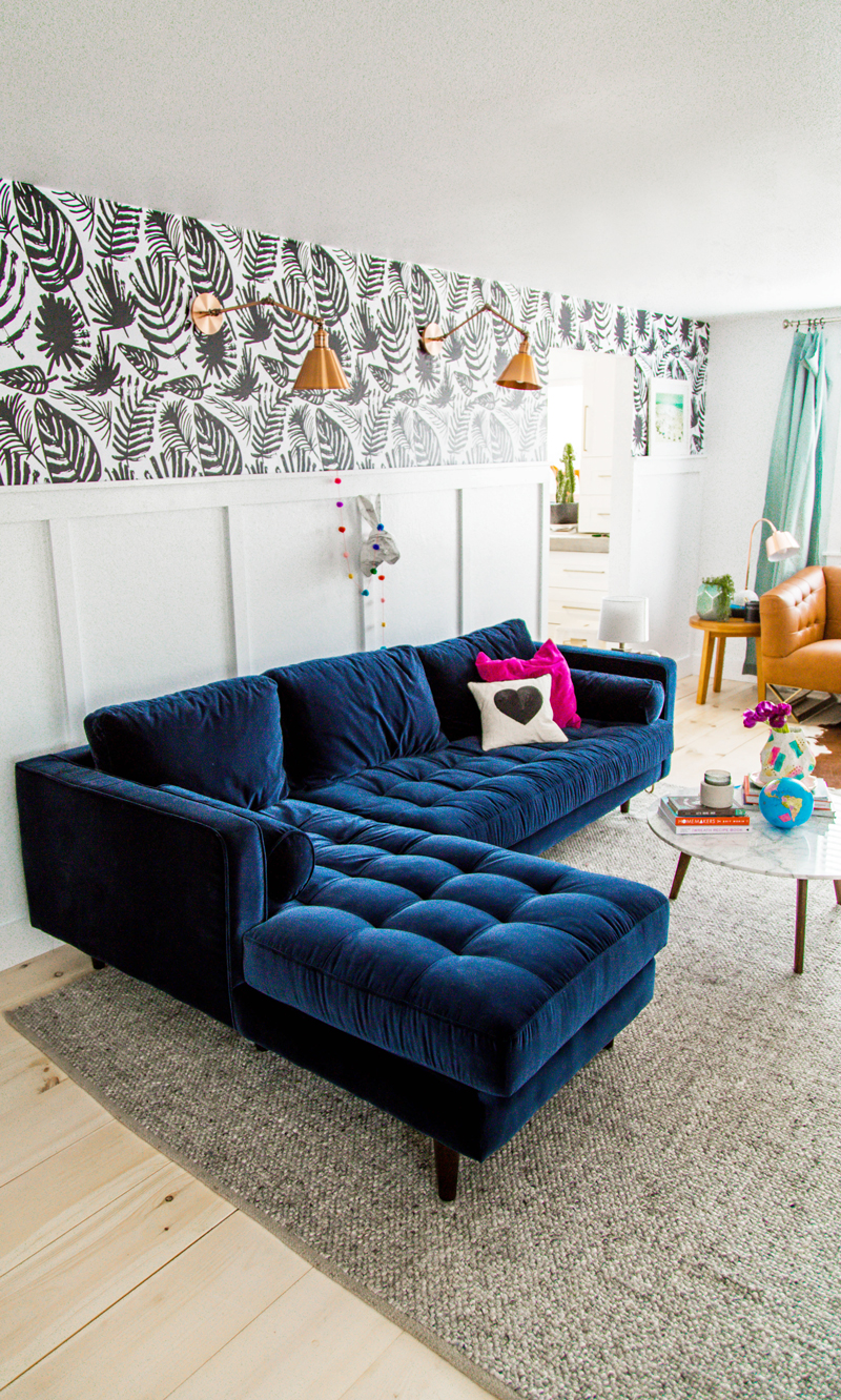 A modern living room with a dark blue velvet sectional sofa, patterned wallpaper, wall lamps, a round coffee table, and a gray textured rug.