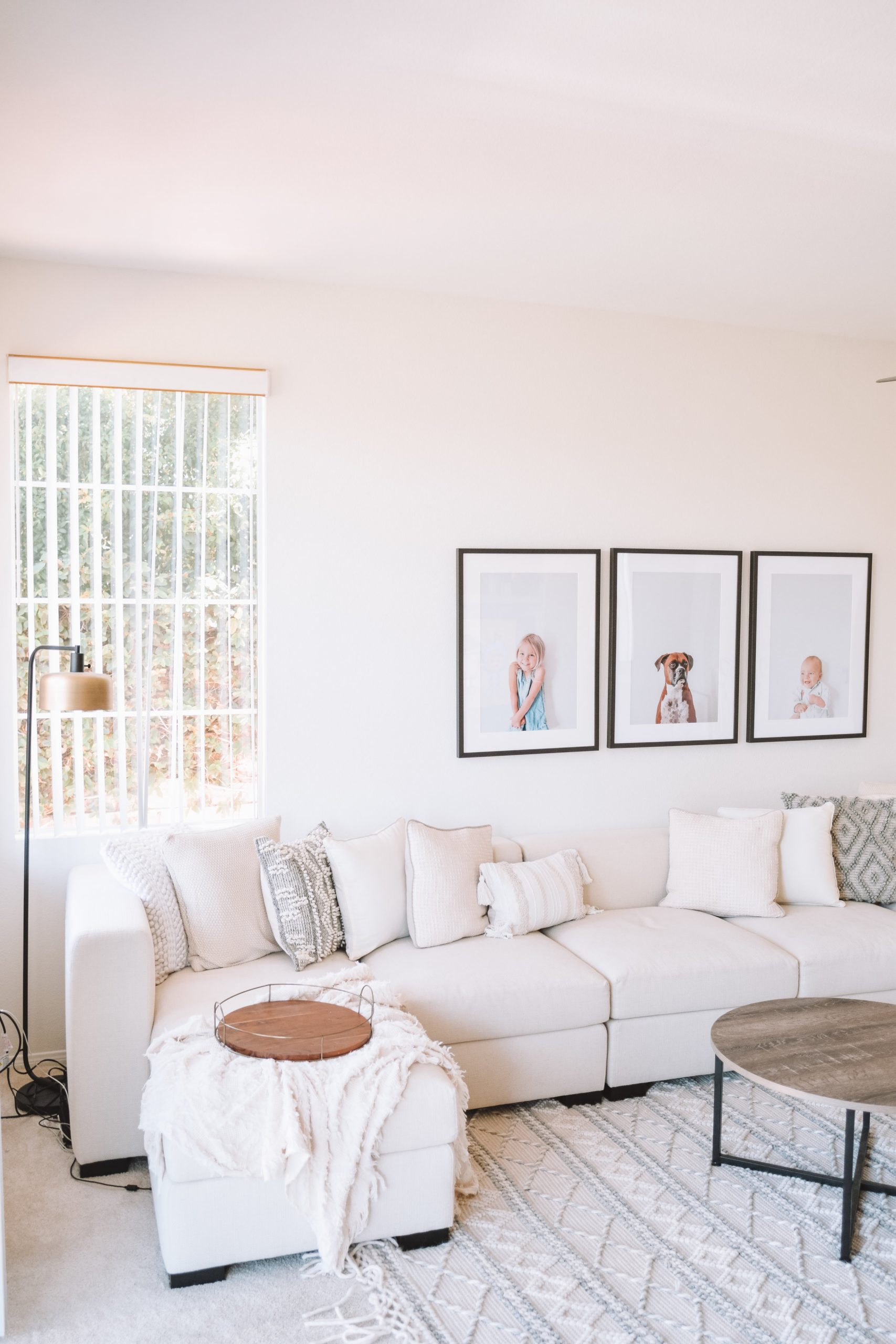 Bright living room with a white sectional sofa, textured pillows, a floor lamp, round coffee tables, and three framed photos on the wall above the couch.