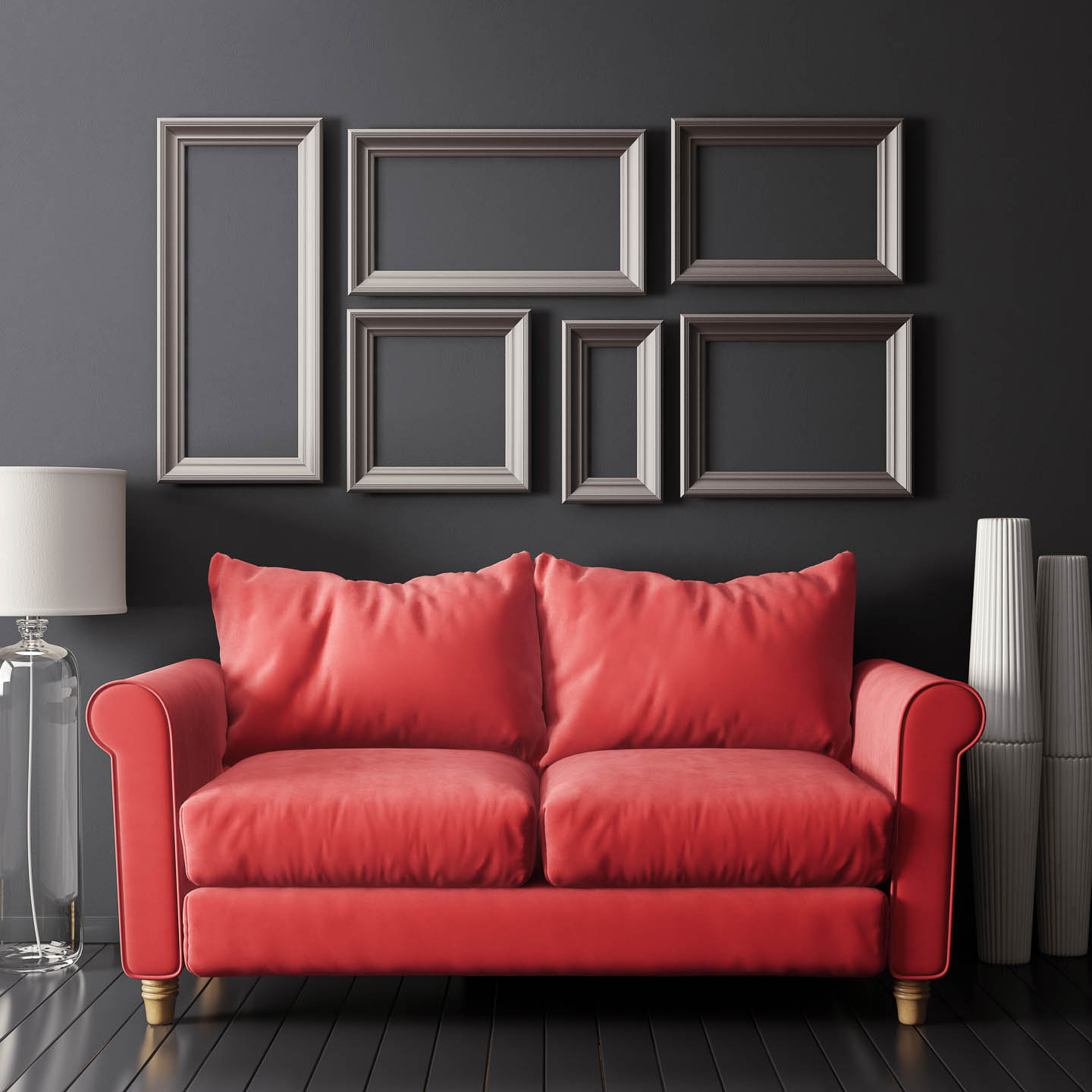 A red sofa sits in front of a dark wall with six empty picture frames; a white lamp and stacked vases are placed to the sides.