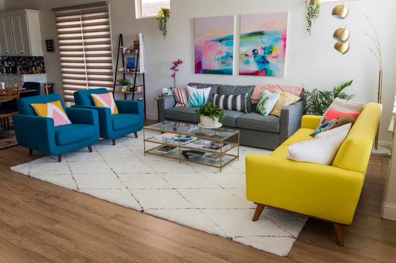 A modern living room with a gray sofa, three teal armchairs, a yellow couch, a glass coffee table, abstract wall art, and plants on a white rug with wooden flooring.