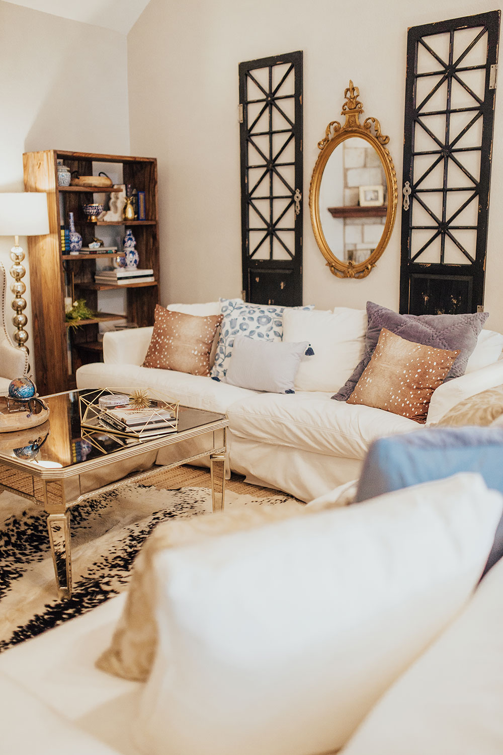 A living room with white sofas, decorative pillows, a mirrored coffee table, a wooden bookshelf, and a gold-framed mirror between two black lattice panels on the wall.