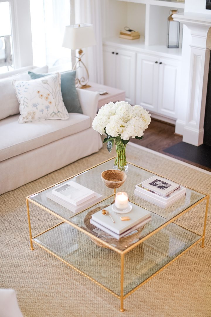 A square glass coffee table decorated with books, some flowers, a bowl and a candle.