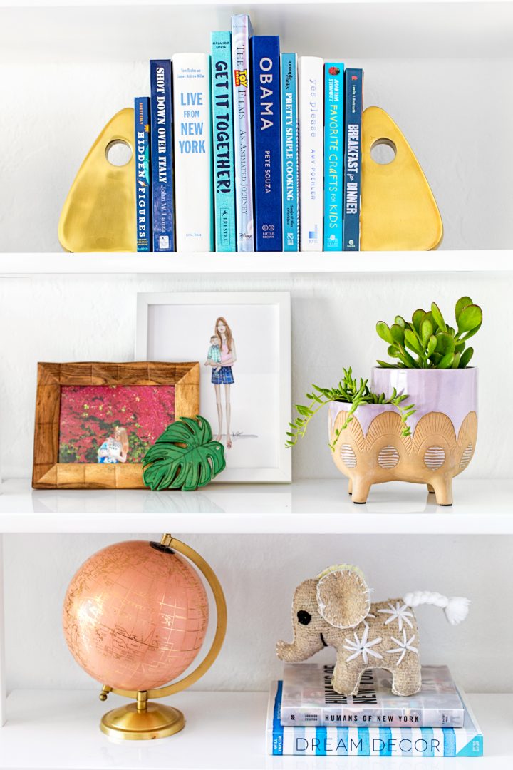 Modern bookends at either end of a row of blue hued books on a white bookshelf with frames, plants and a globe