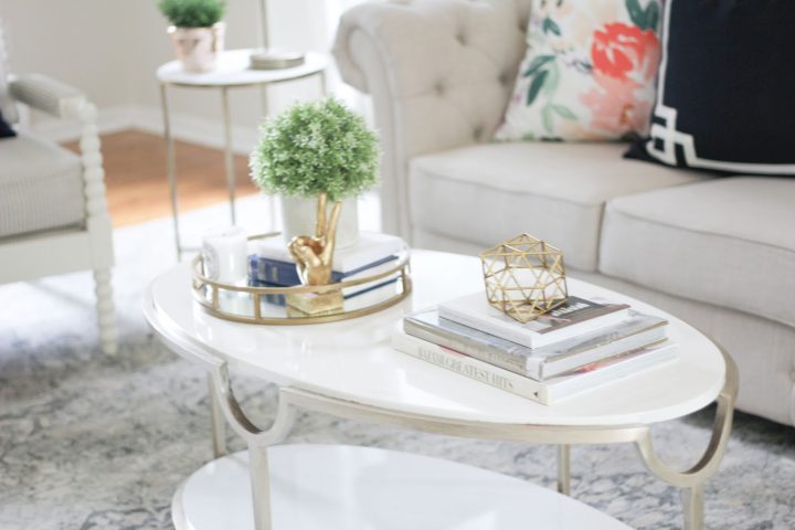 Faux potted plant, books and tray on an oval coffee table, with other gold accessories. 