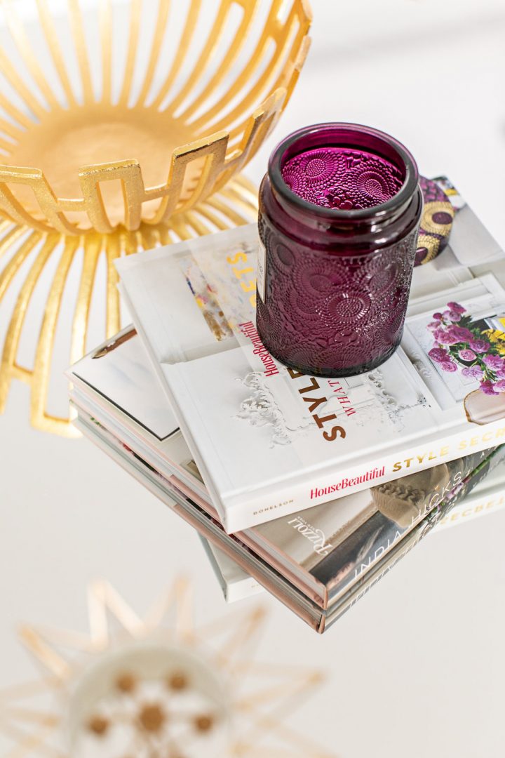 Intricate purple glass candle jar on a stack of coffee table books beside a gold bowl.