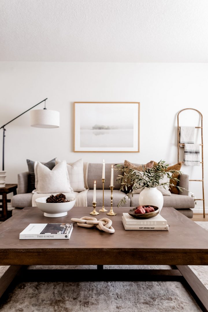 Gold candle holders with white tapered candlesticks, bowls, books, a vase, and other accessories on a square wooden coffee table.
