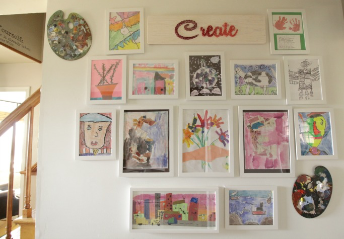 Kid's paintings and drawings displayed in white photo frames on a wall. 