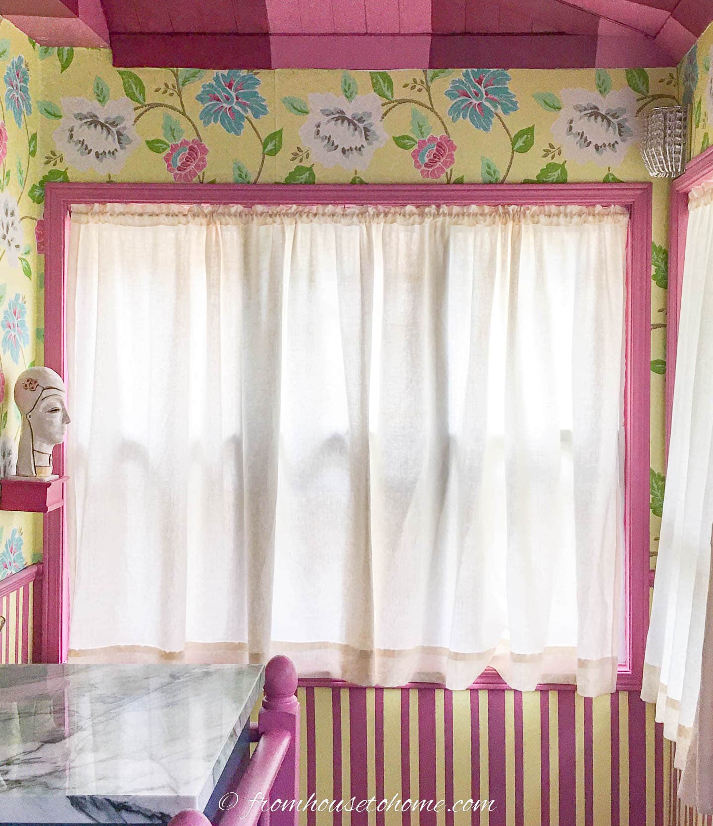 White curtains hung in a window surrounded by bright yellow, pink and blue wallpaper on the wall