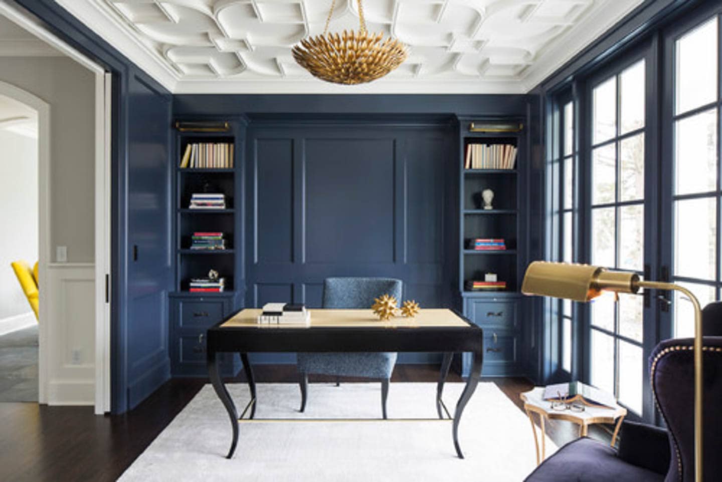 Home office with dark blue walls and shelves, a large window, white ceiling and area rug, and black desk