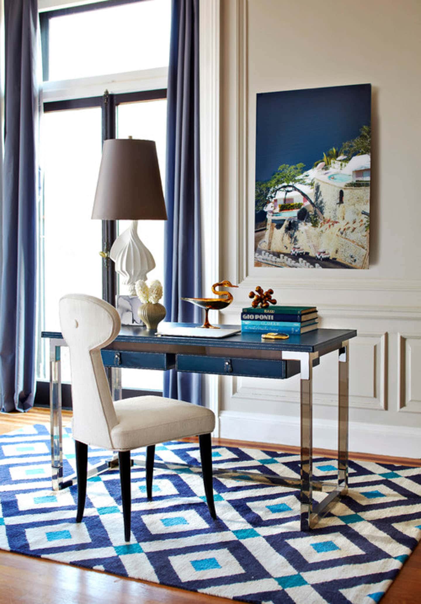 Small blue desk on a blue and white geometric area rug in a white room with dark blue curtains