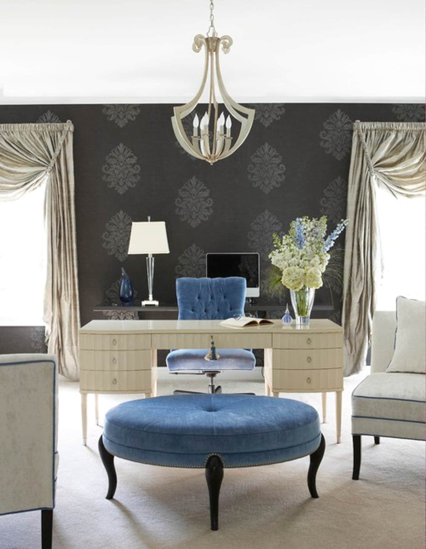 Sophisticated home office with dark gray walls, cream desk and curtains, and a blue velvet chair and ottoman