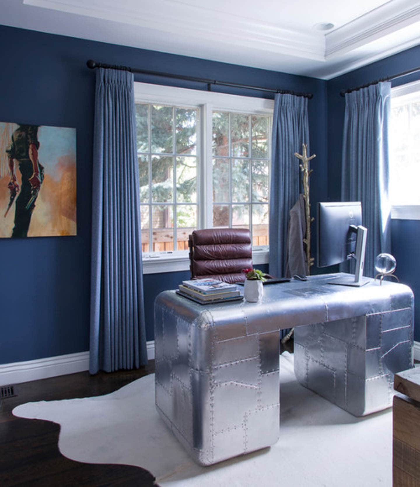 Home office with dark blue walls and curtains and a ilver riveted desk in front of a window
