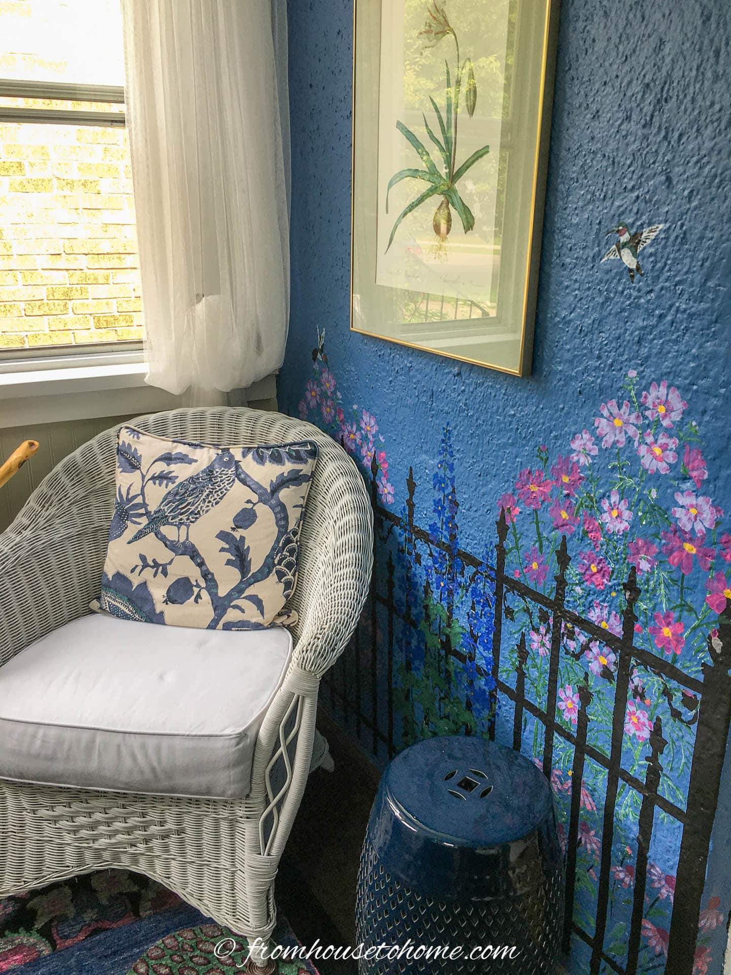 small blue and white front porch with a white wicker chair, blue ceramic stool and a blue wall with a stenciled garden