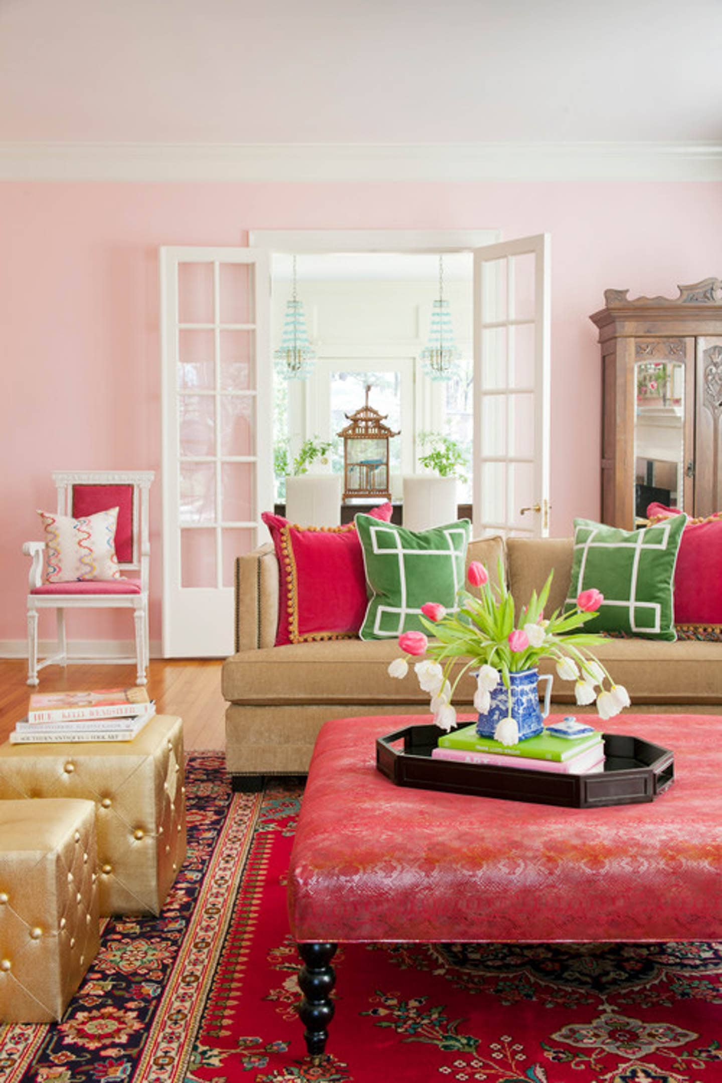 Living room with pink walls, tan sofa, red and green throw pillows and a red ottoman