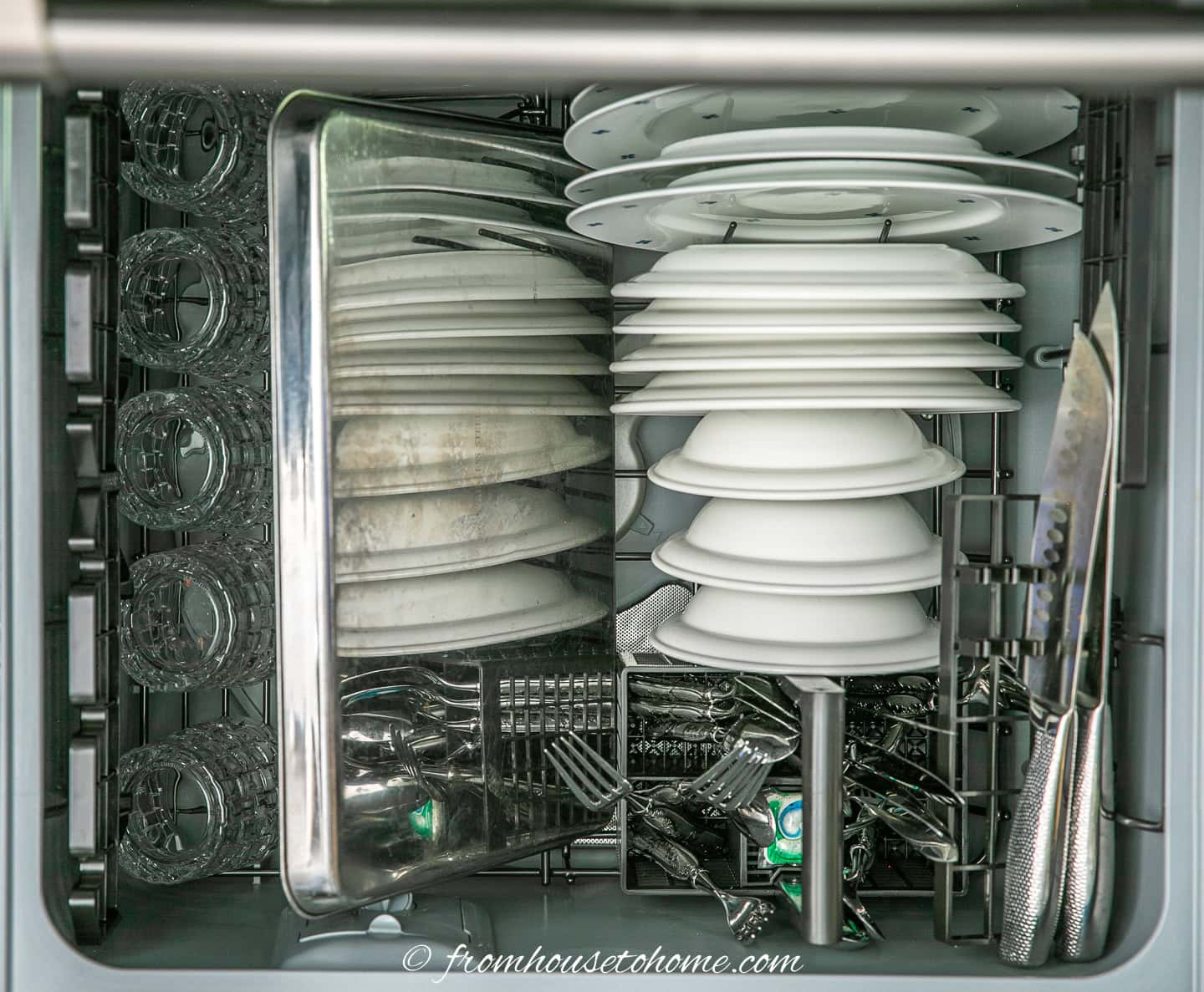 a dishwasher drawer filled with plates, bowls, glasses, cutlery and a baking sheet