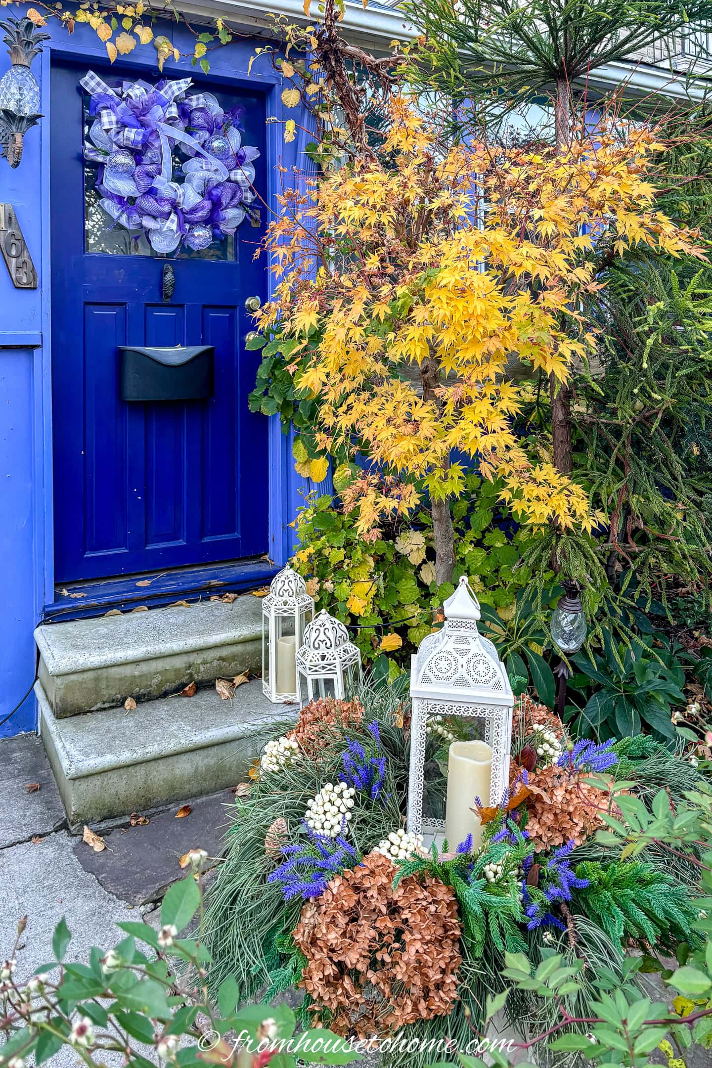 lavender and white deco mesh Christmas wreath hanging on the front door of a house with a purple and white winter planter in front of it and a yellow Japanese maple beside it