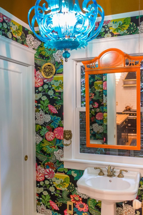 A turquoise chandelier hung in a small bathroom with floral wallpaper and an orange framed mirror