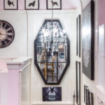 A chandelier hangs over a stairwell, reflected in a large octagonal mirror surrounded by framed art and silhouettes; a large clock and bookshelves are visible on the left.