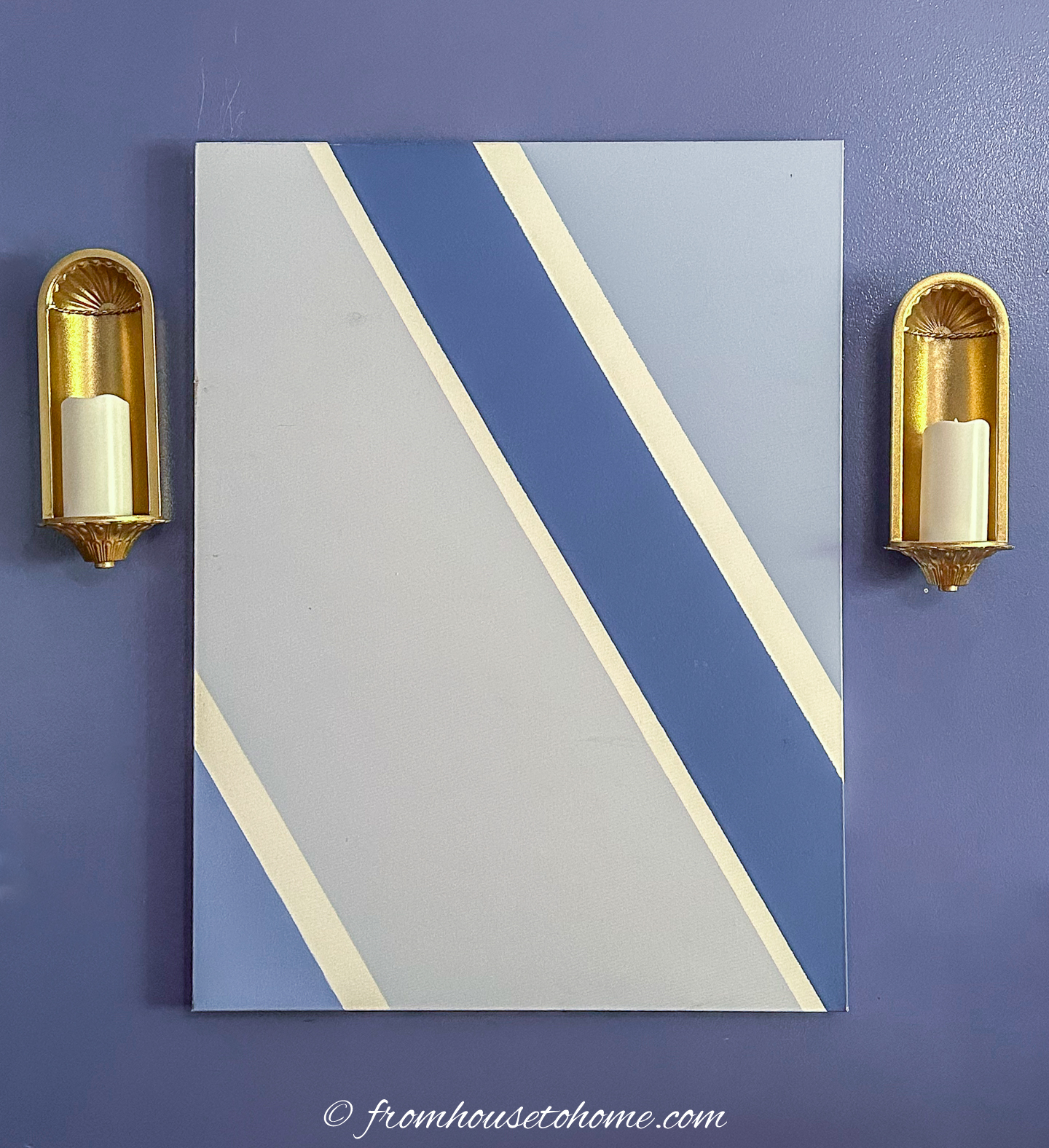 A geometric painting with diagonal beige, blue, and cream stripes on a gray background, flanked by two gold wall sconces with candles.