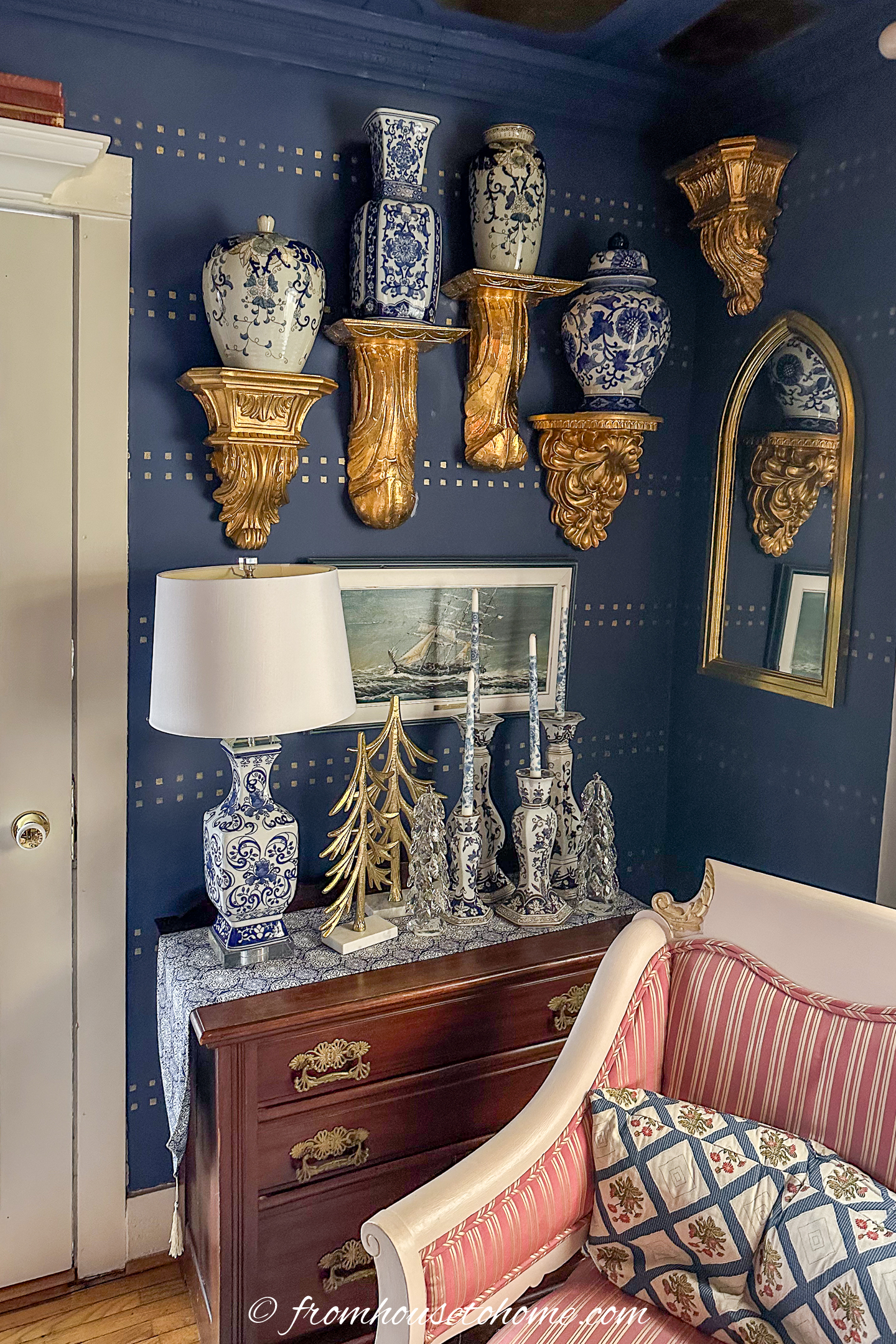 A traditional living room corner with blue walls, gold shelves holding blue-and-white vases, a mirror, table lamp, silver decor, and a pink-striped sofa.