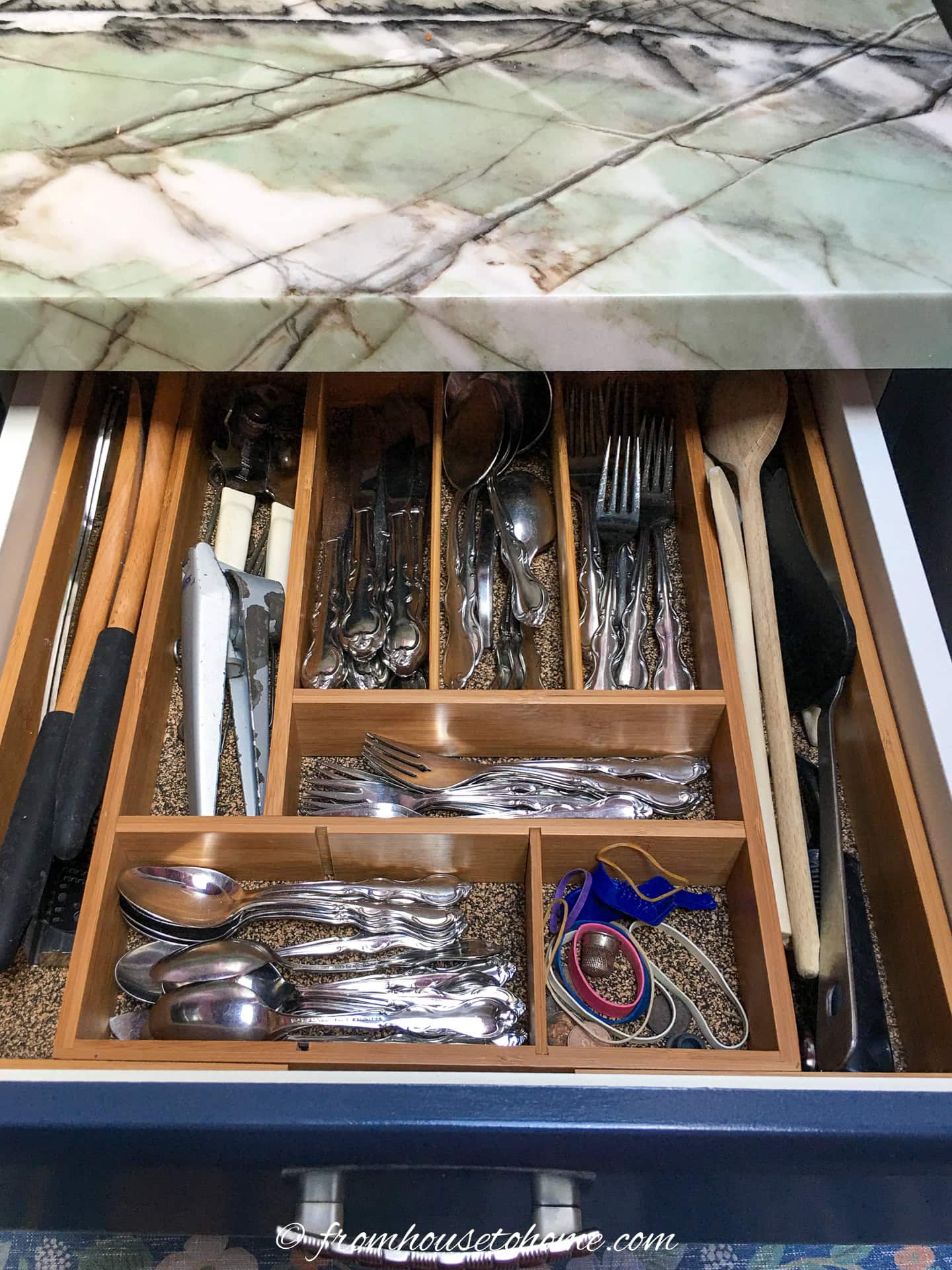 A kitchen drawer organized with wooden dividers holding silverware, utensils, knives, and miscellaneous items under a marble countertop.