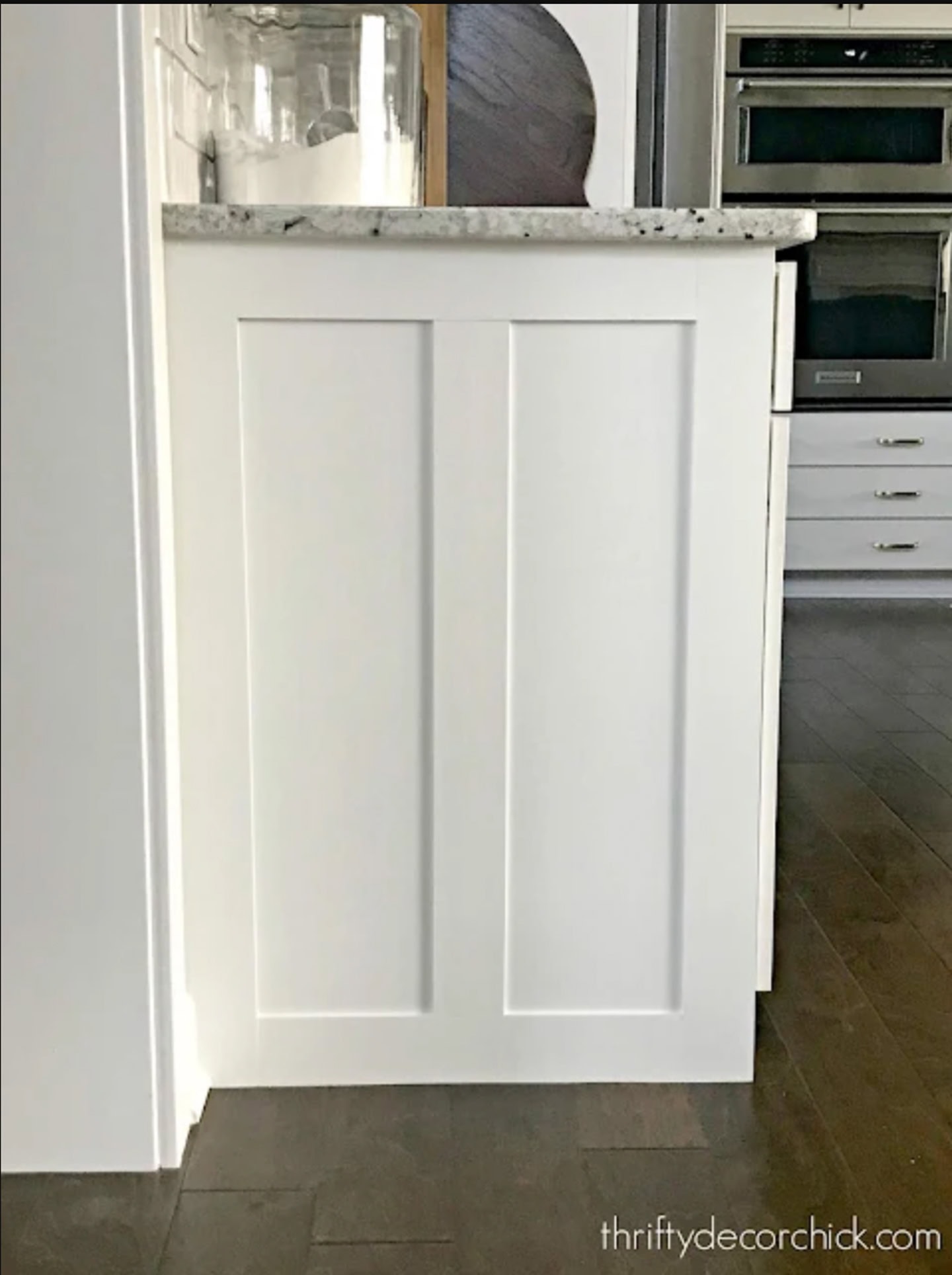 A white shaker-style kitchen cabinet end panel with a marble countertop, set against a dark wood floor.