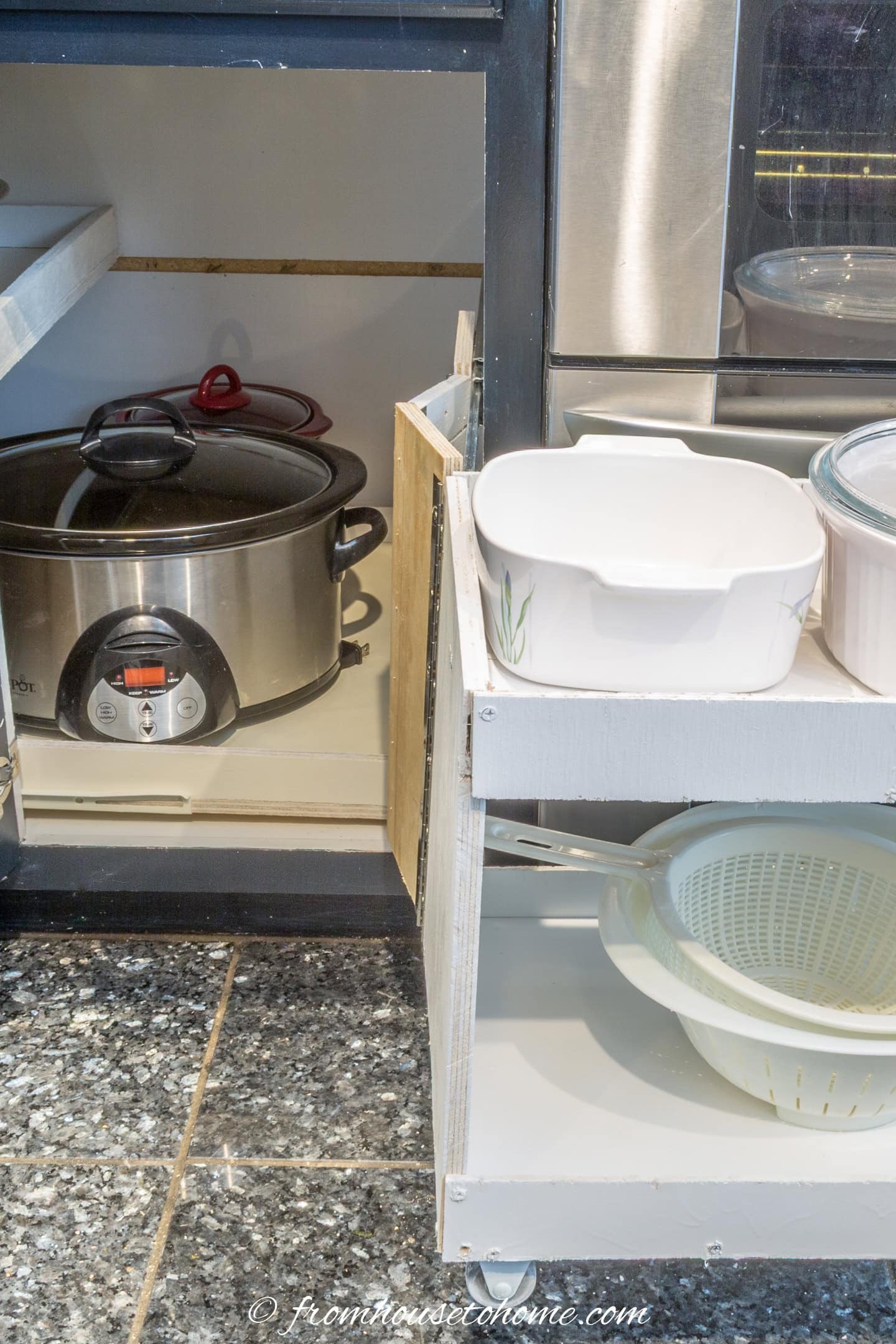 A kitchen cabinet with a slow cooker inside and a pull-out shelf holding a white bowl, a lidded dish, and a colander.