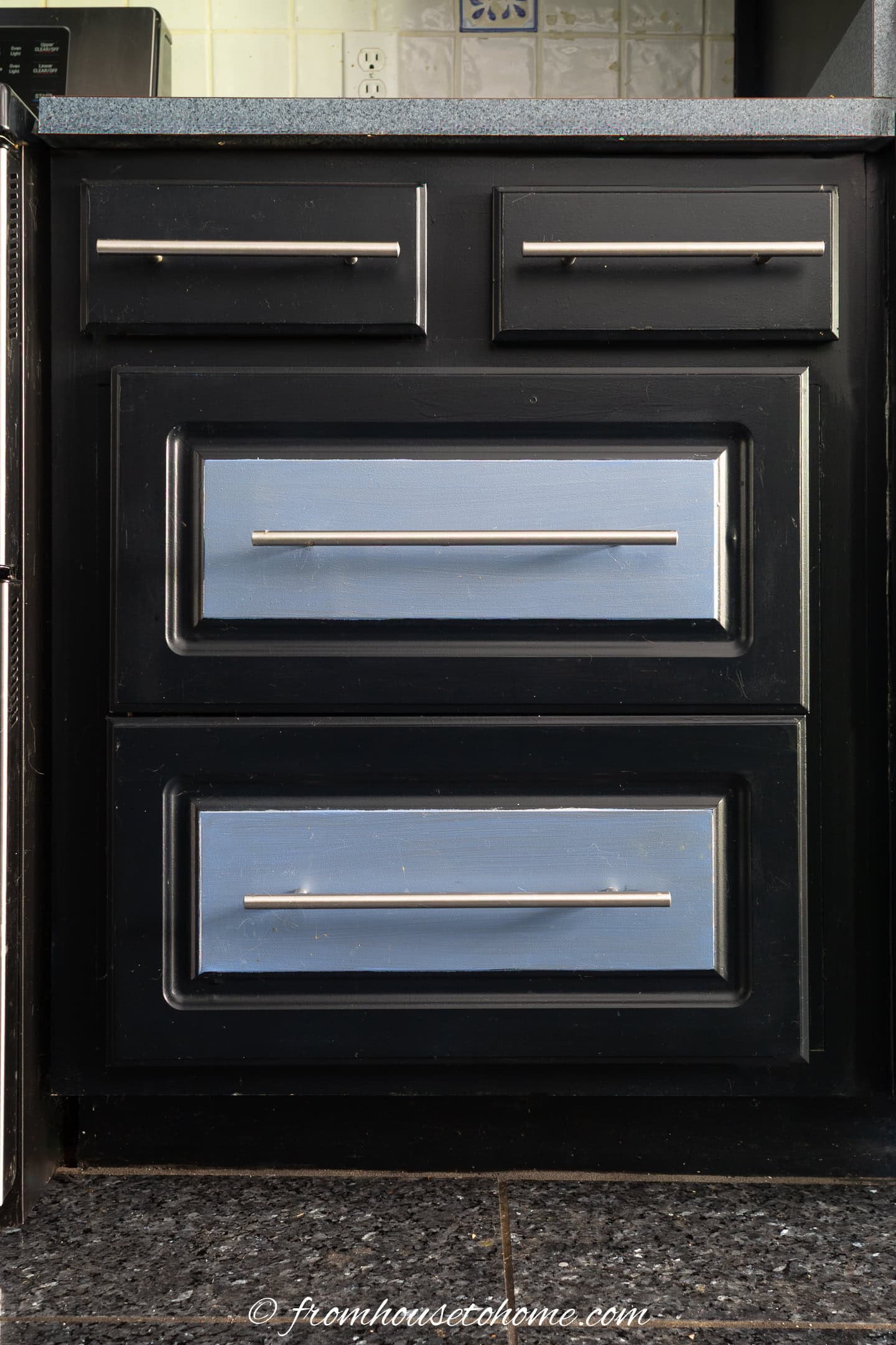 A close-up view of black kitchen cabinets with drawers, silver handles and light blue drawer fronts, set against a tiled backsplash and dark countertop.