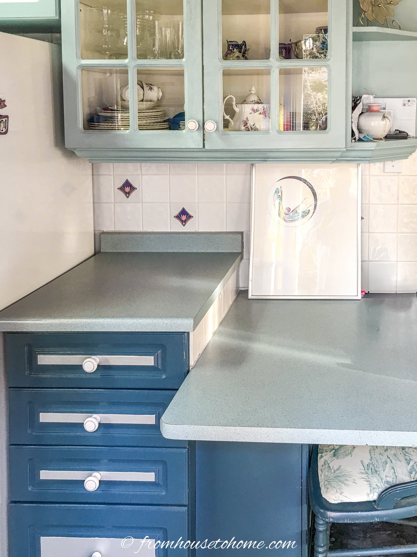 A kitchen corner with blue cabinets, glass-front upper doors, vintage teapots, tiled backsplash, and a framed picture displayed on the countertop.