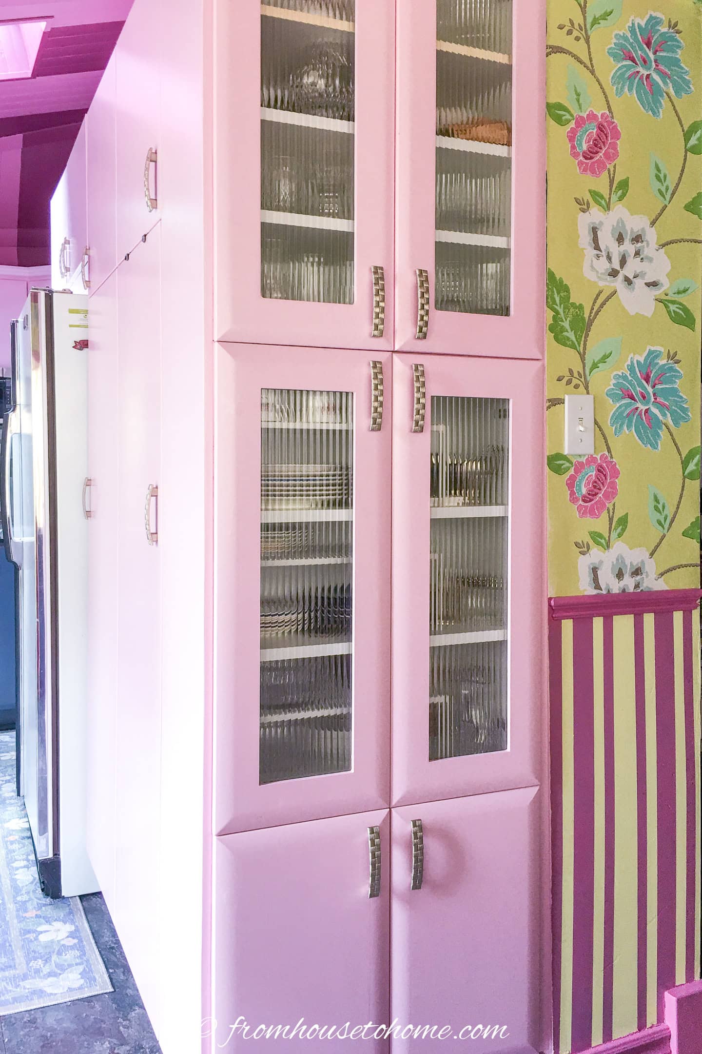 Tall pink kitchen cabinets with glass-paneled doors are shown next to a floral wallpapered wall and a striped wainscoting. Shelves with dishes are visible through the glass.