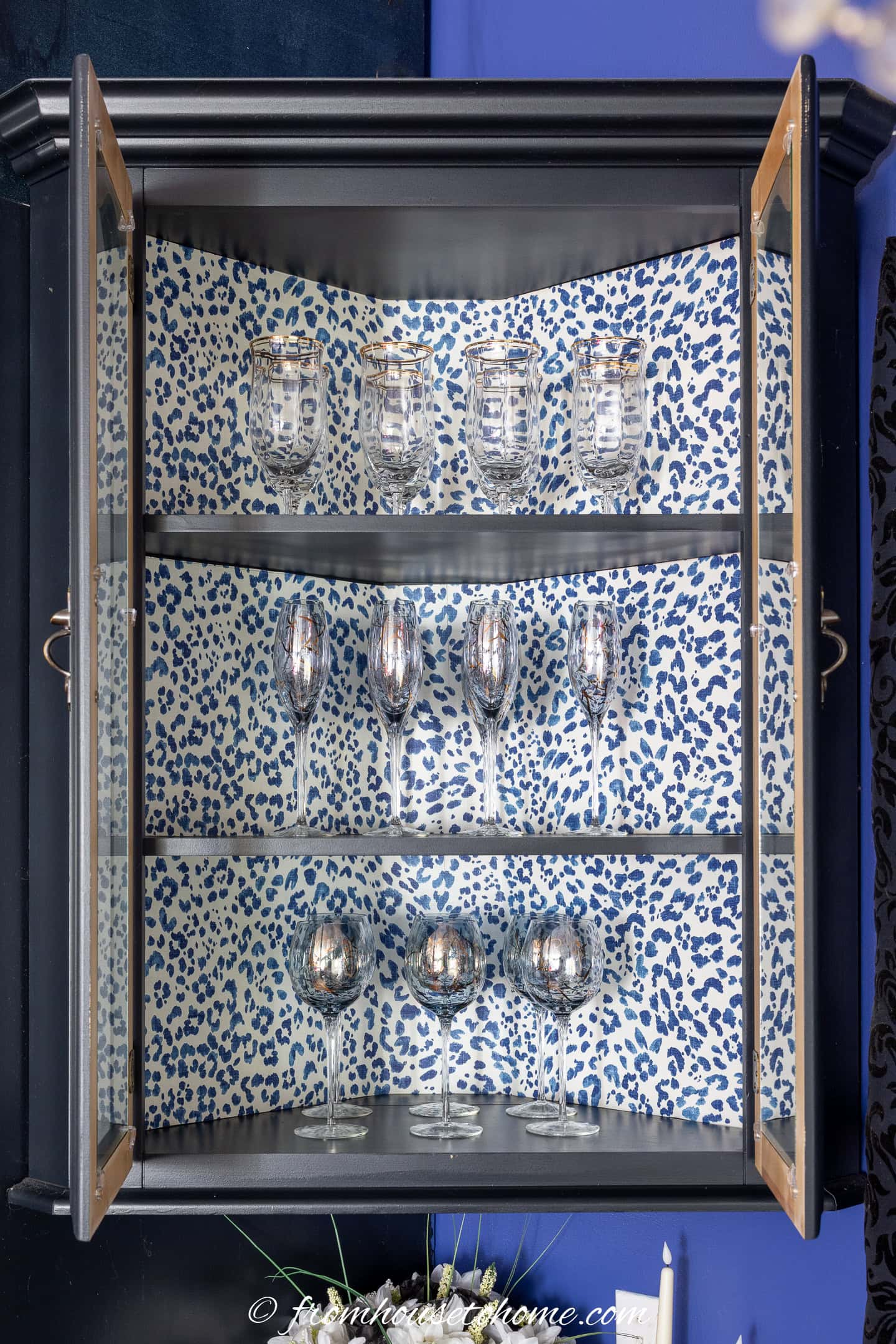 A corner cabinet with glass doors displays silver goblets and wine glasses on two shelves, with a blue and white animal print backing.