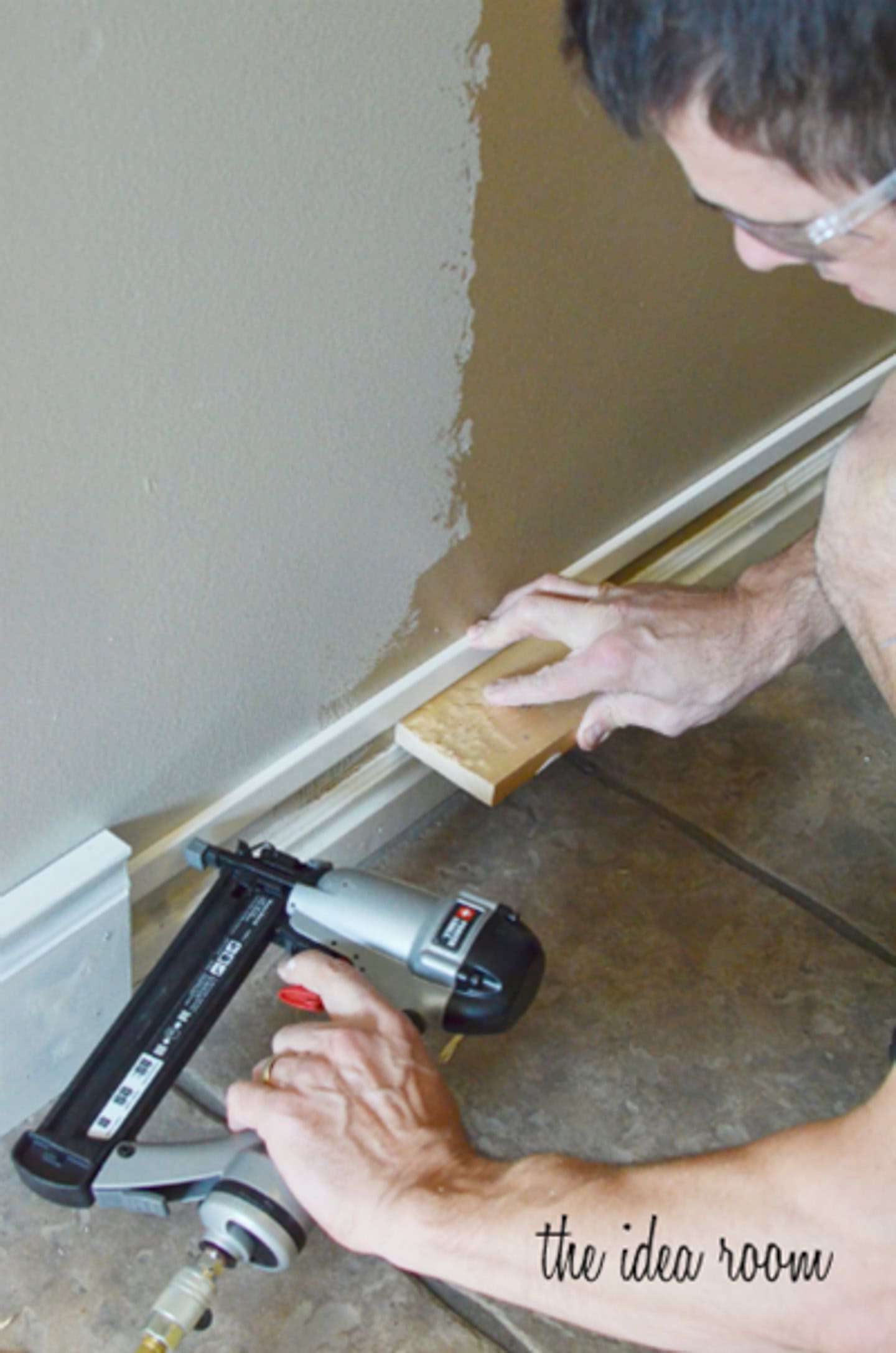 A person uses a sanding block to smooth baseboards while holding a pneumatic nail gun on a tiled floor near a painted wall.