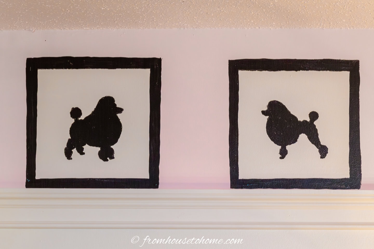 Two black poodle silhouettes are painted on white canvases with black borders, displayed side by side on a light pink wall above a white shelf.
