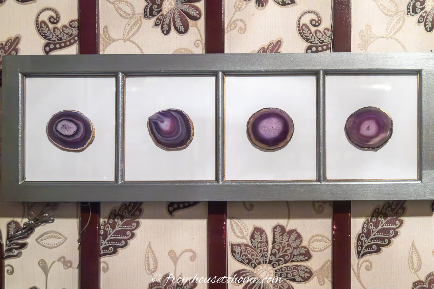 A framed display with four slices of purple agate, each set in a separate section, hangs on a wall with a floral and stripe pattern.