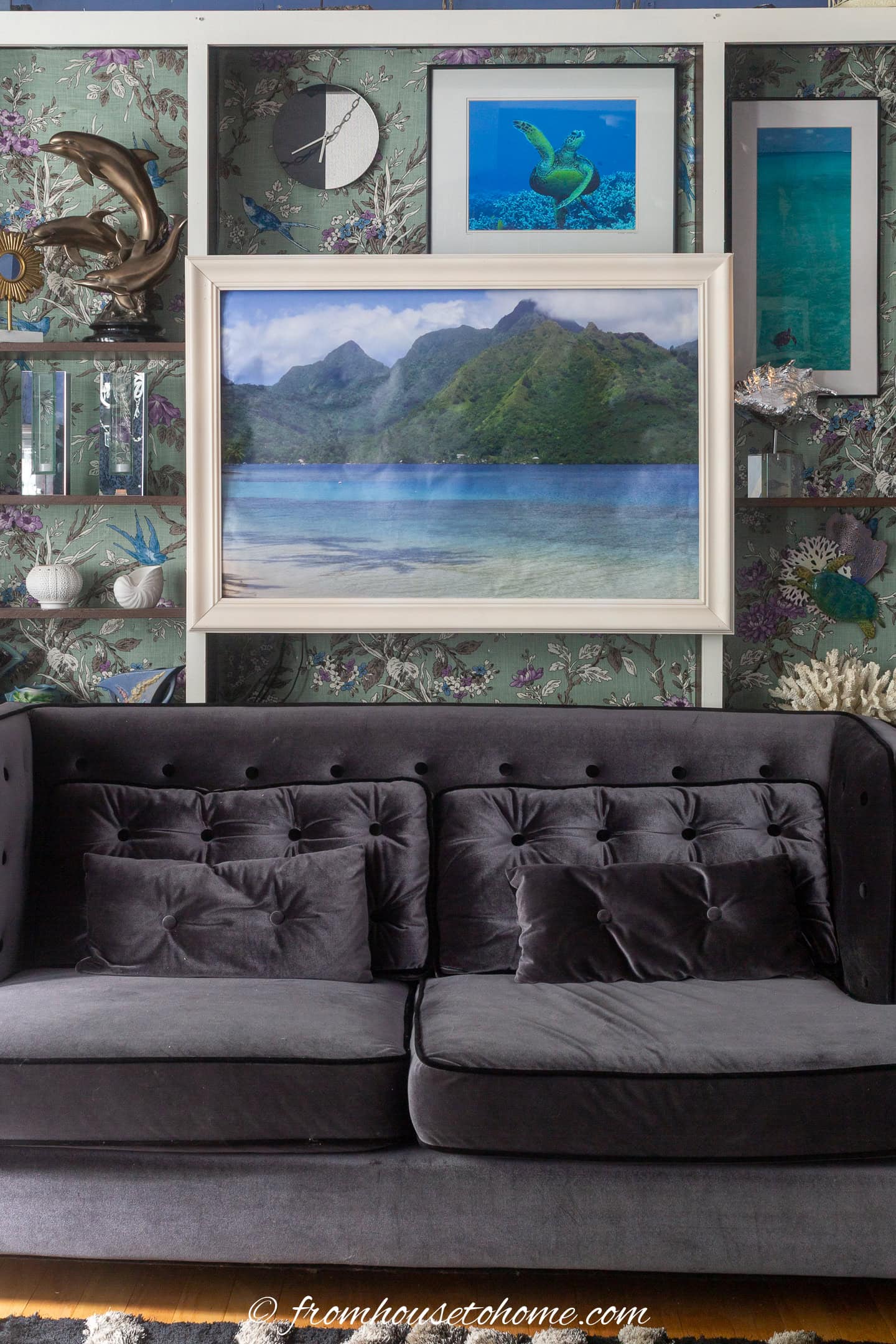 A gray velvet sofa sits against a wall decorated with framed landscape and sea life art, floral wallpaper, and various decorative objects.