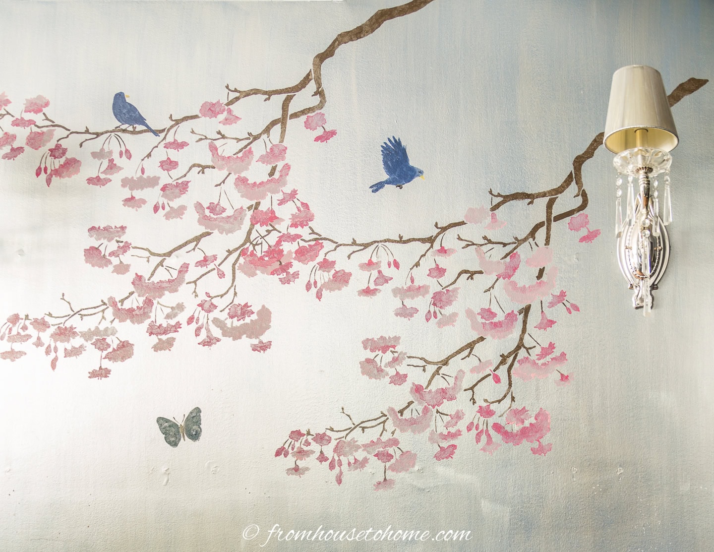 A wall mural features cherry blossom branches with pink flowers, two blue birds, a butterfly, and a wall-mounted glass sconce on the right.