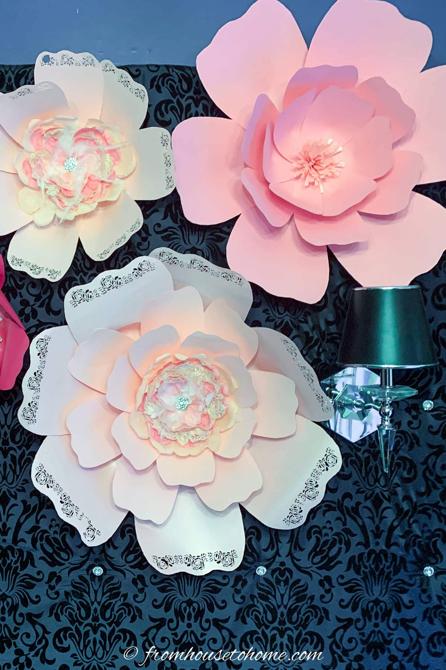Large pink paper flowers with decorative edges are mounted on a black patterned wall next to a black wall lamp with a crystal base.