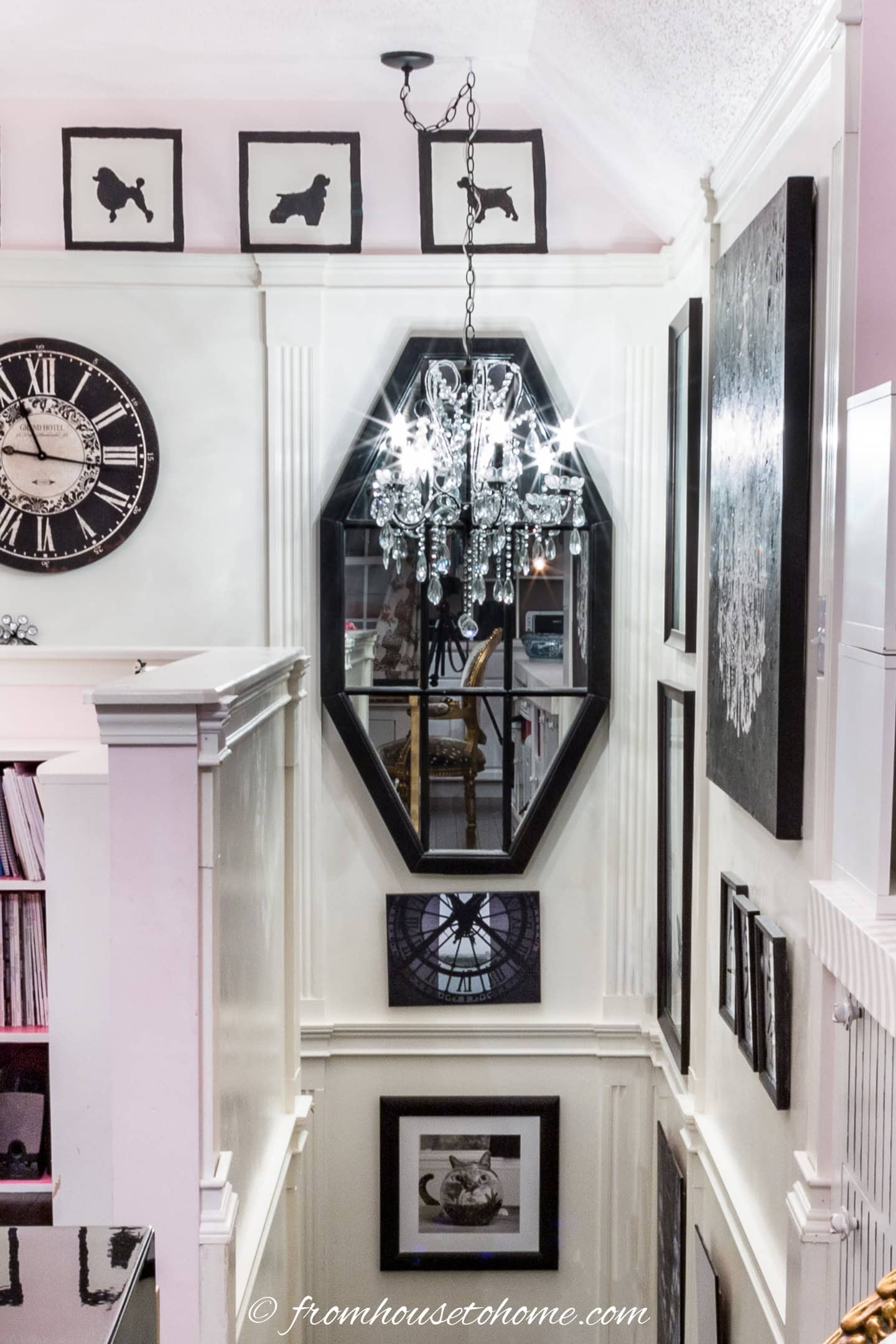 A chandelier hangs over a stairwell, reflected in a large octagonal mirror surrounded by framed art and silhouettes; a large clock and bookshelves are visible on the left.