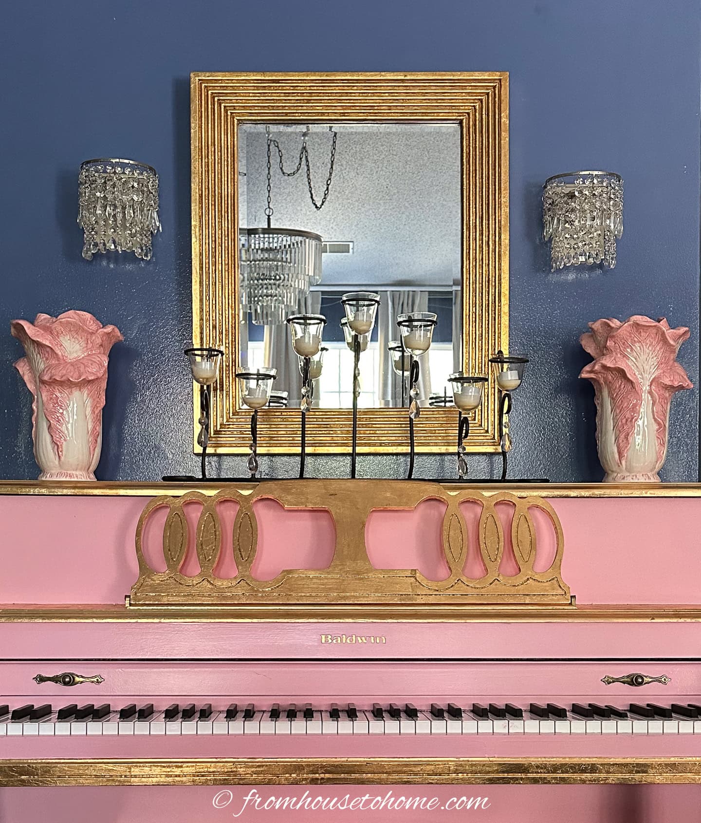 A pink and gold Baldwin piano sits against a blue wall, topped with a gold candelabra, pink vases, and a gold-framed mirror flanked by crystal wall sconces.
